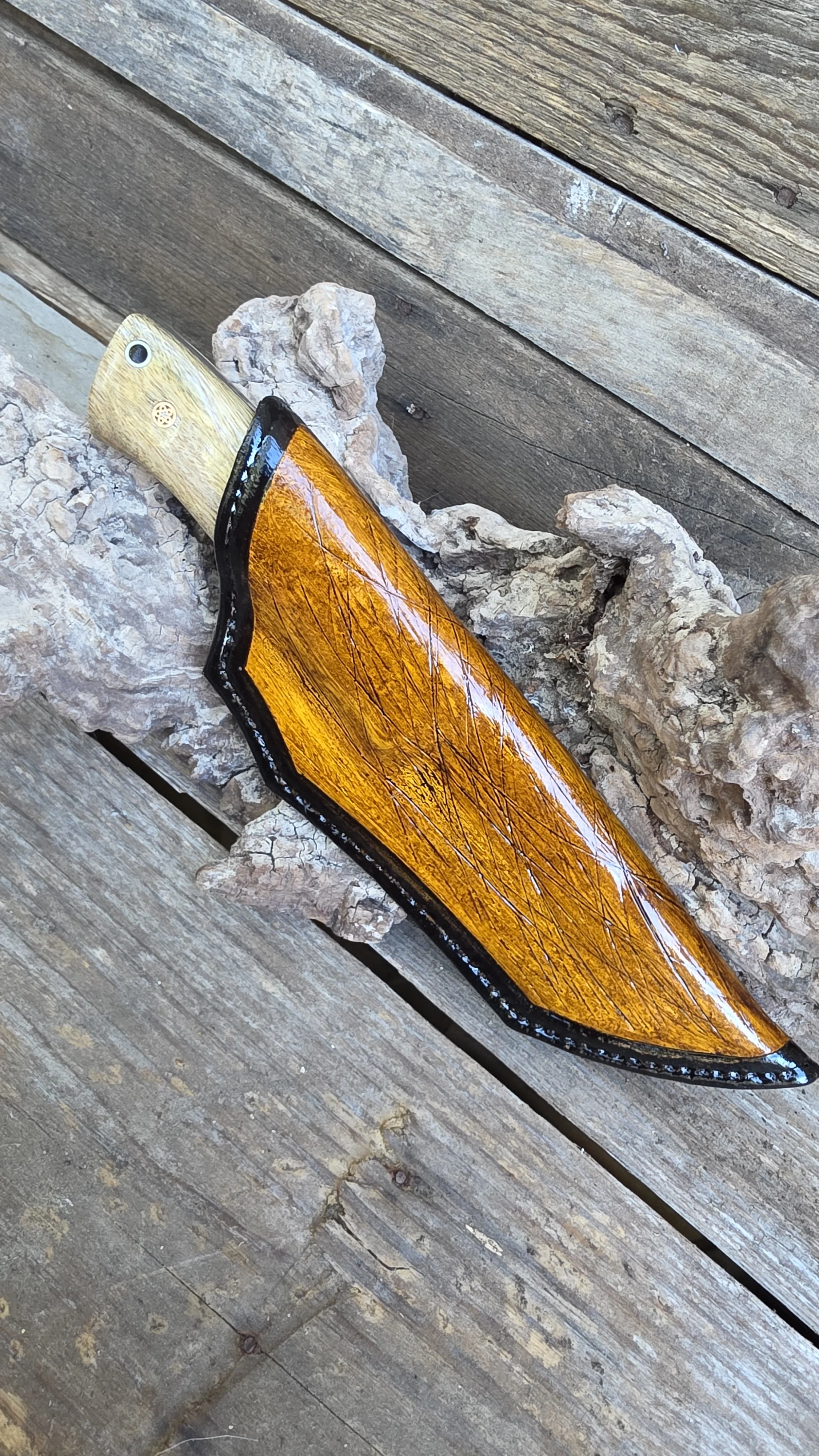 A hunting knife with a light wooden handle and a black-edged blade, resting on a piece of driftwood on a weathered wooden surface.
