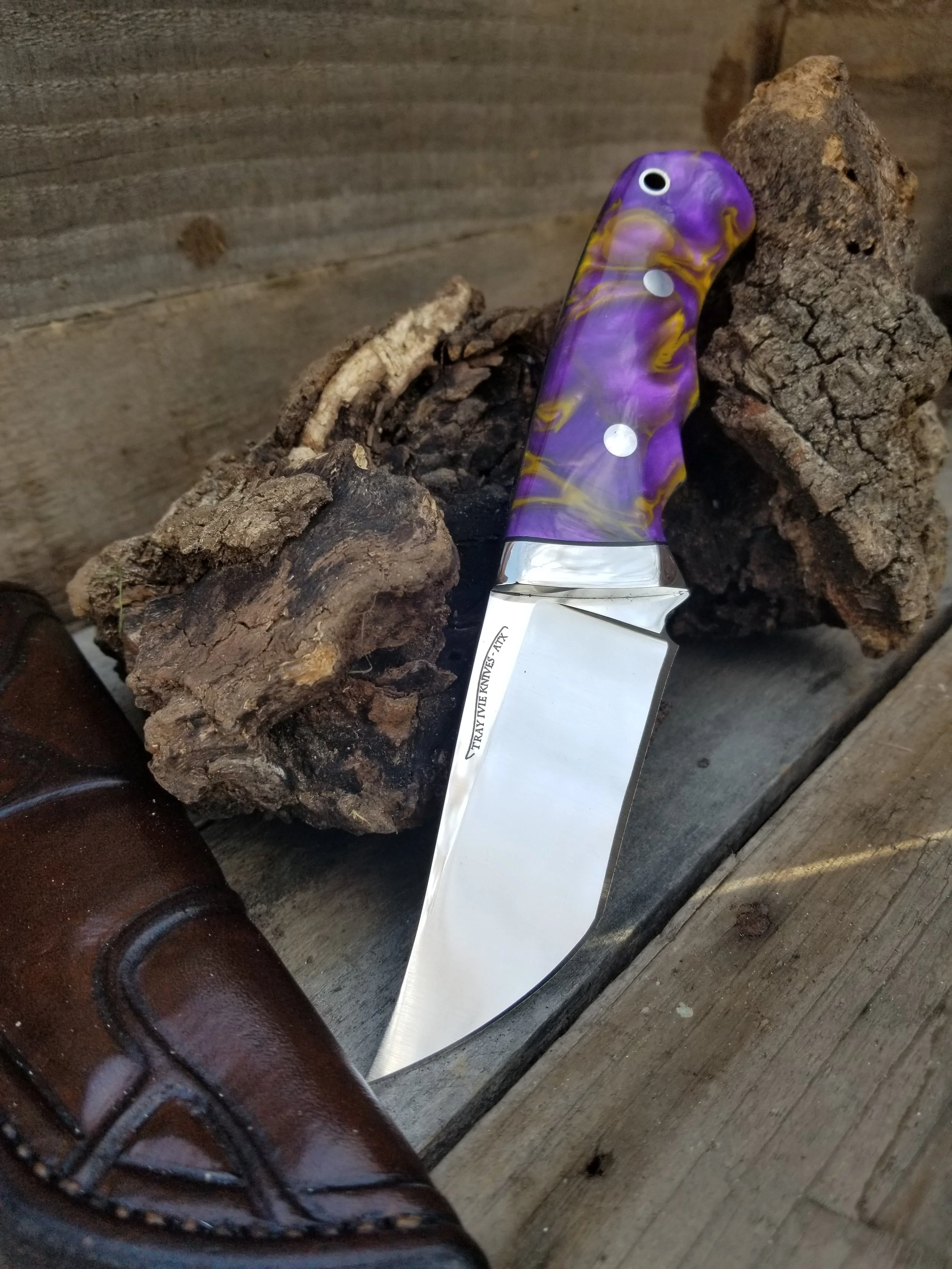 A kitchen knife with a purple and yellow swirled handle, placed on a piece of bark with a wooden wall in the background.