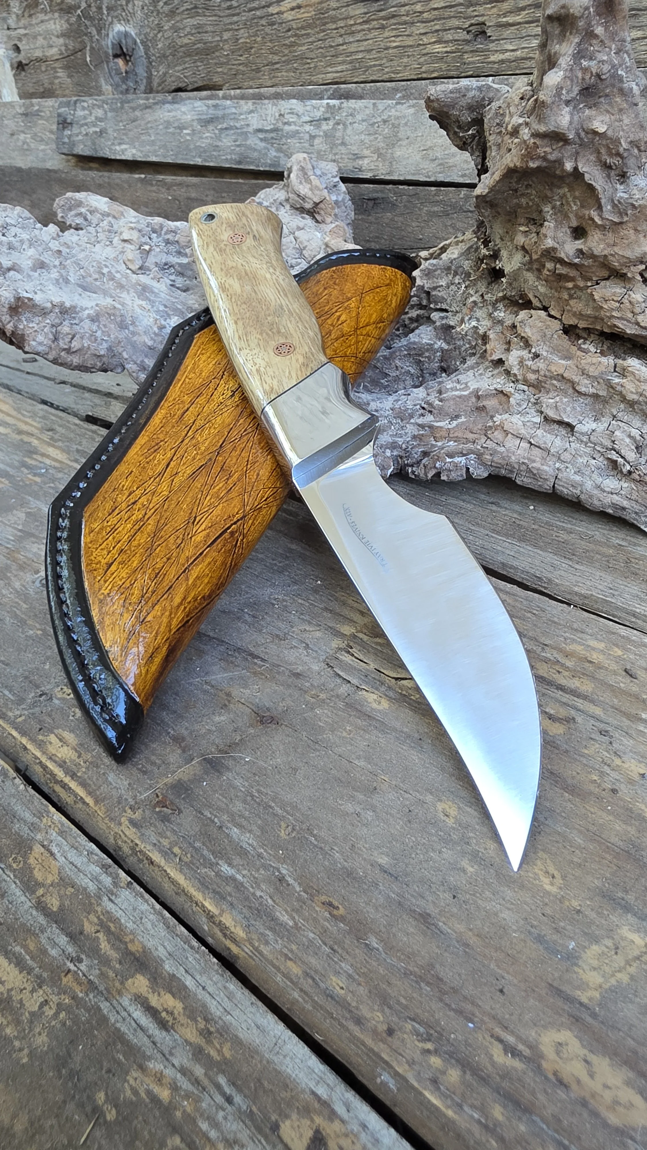 Close-up of a handmade knife with a stainless steel blade and a wooden handle, resting on a rustic wooden surface with some rocks and driftwood in the background.