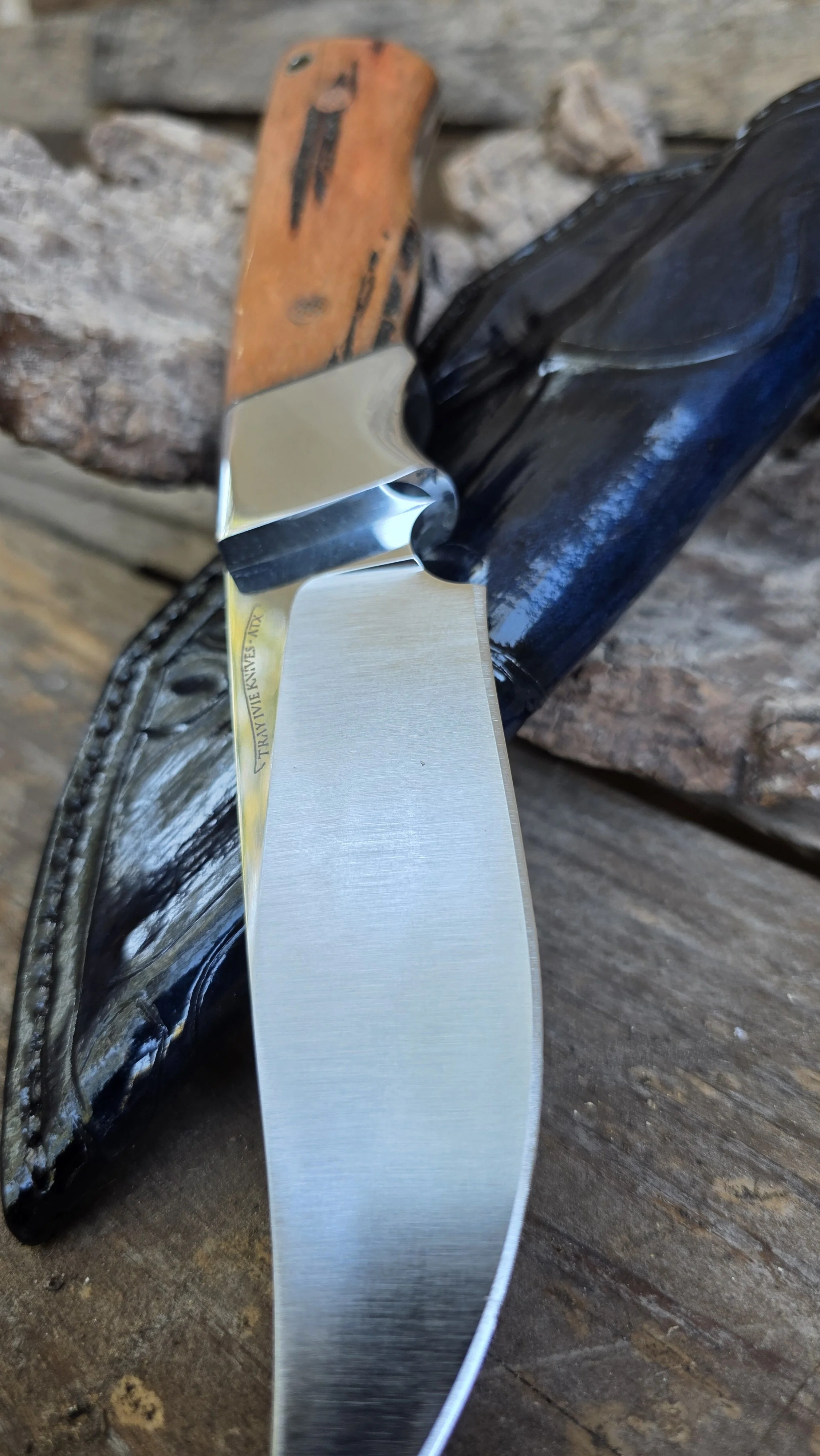 A close-up of a folding pocket knife with a stainless steel blade, resting on a black leather sheath on a wooden surface with some rocks in the background.