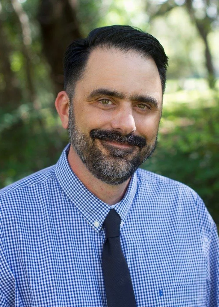 A man with dark hair, a beard, and a mustache wearing a blue checkered shirt and a dark tie, standing outdoors with a blurred green and brown background.