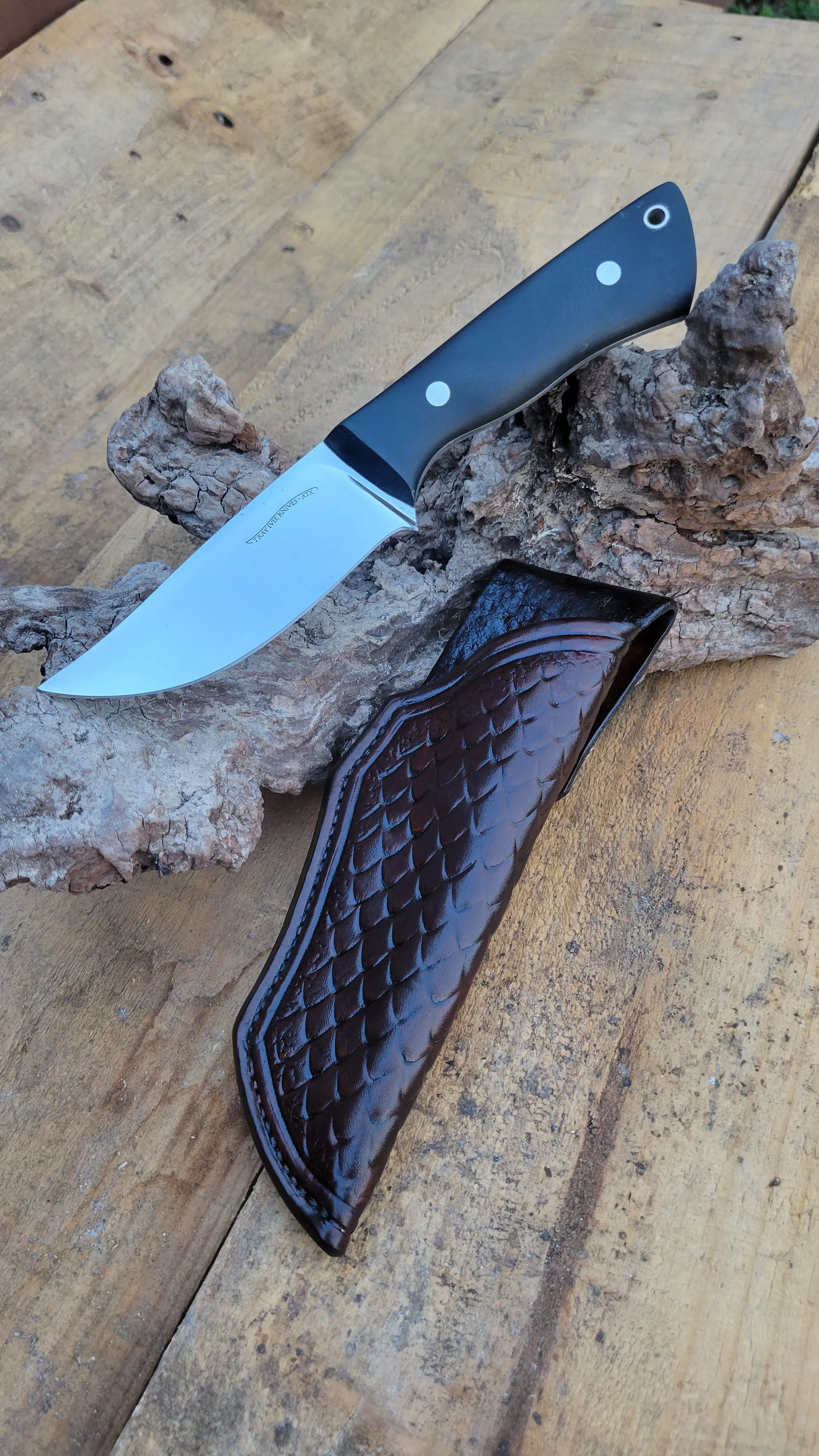 A hunting or utility knife with a black handle and a stainless steel blade resting on a piece of textured wood and a carved brown leather sheath nearby.