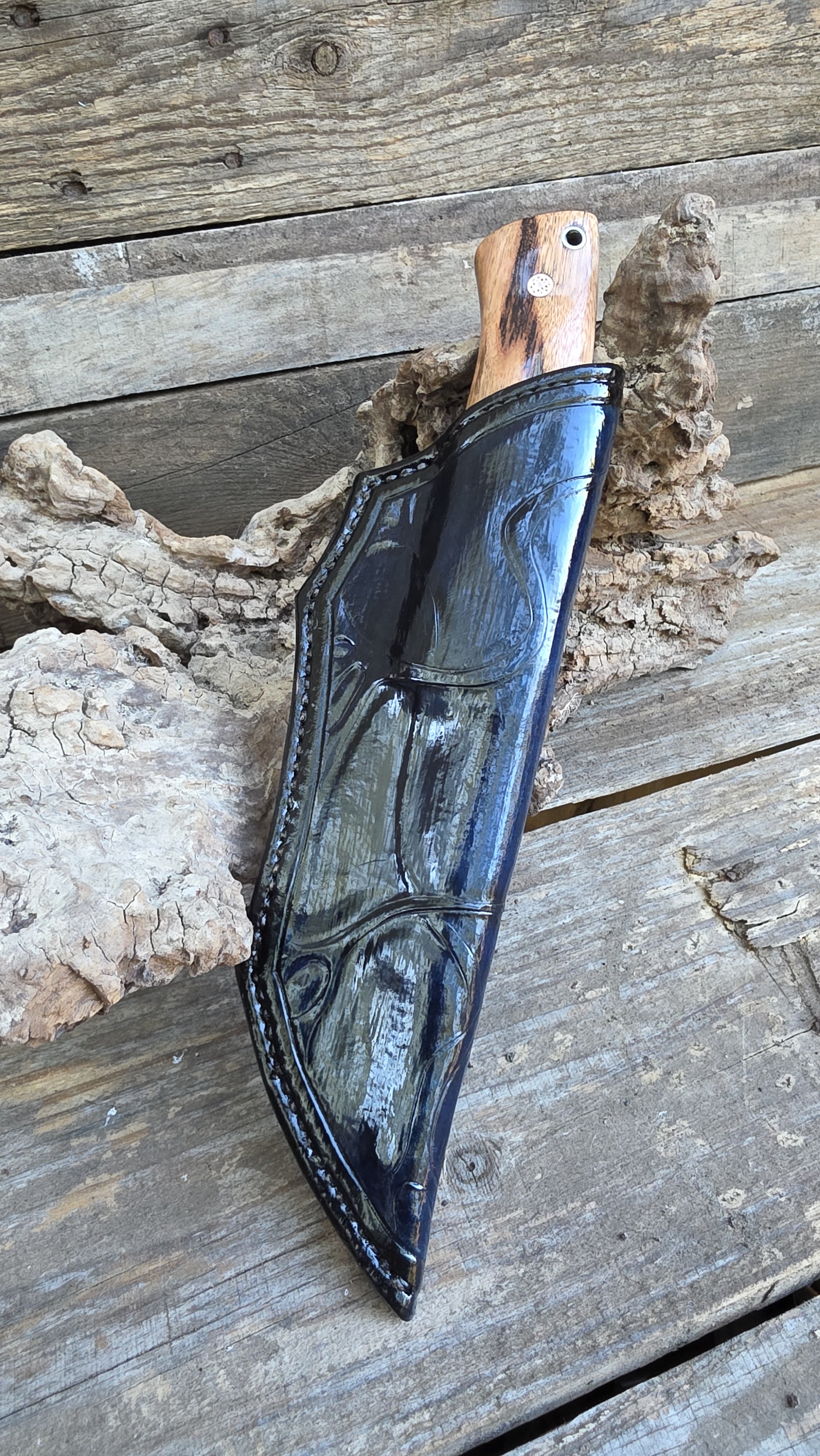 A fishing knife with a wooden handle featuring googly eyes and a white circular sticker, resting in a black leather sheath on a rustic wooden surface with a piece of driftwood.