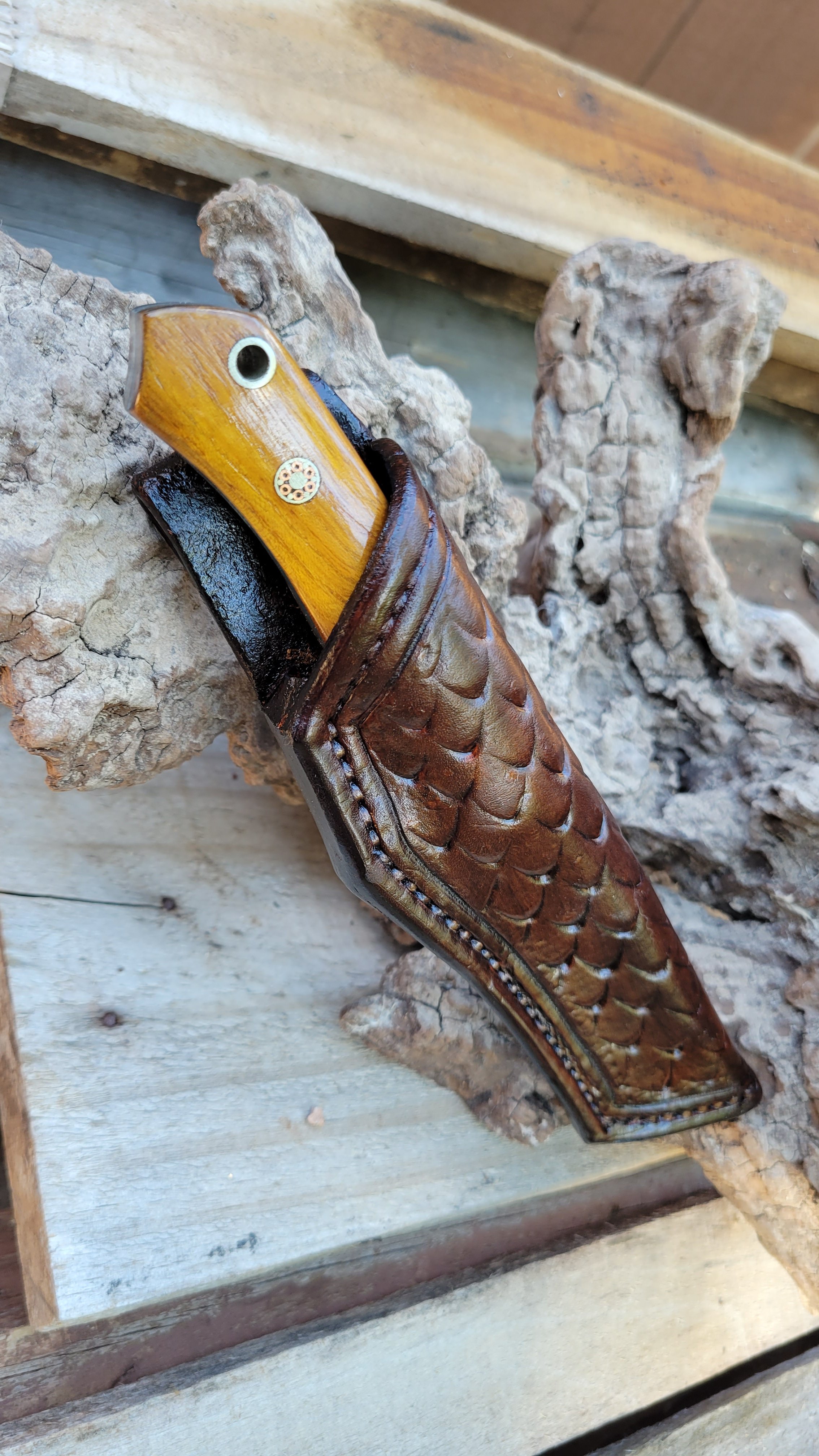 A knife with a wooden handle and a metal eye in it is inside a brown leather holder shaped like a fish, resting on a piece of driftwood or bark.