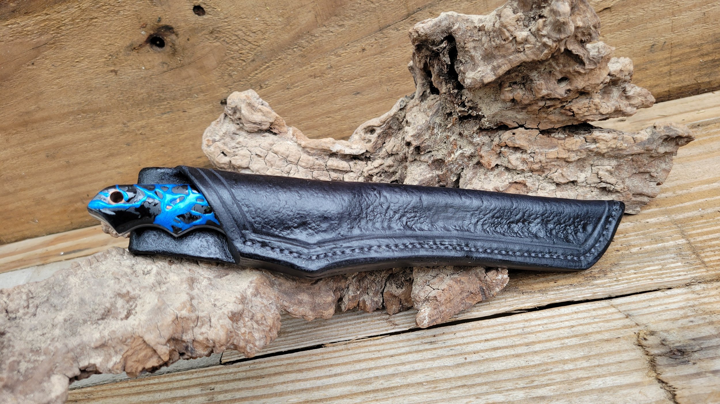 A folding knife with a black textured handle and a blue and black patterned accent, resting on a piece of wood and rocks.