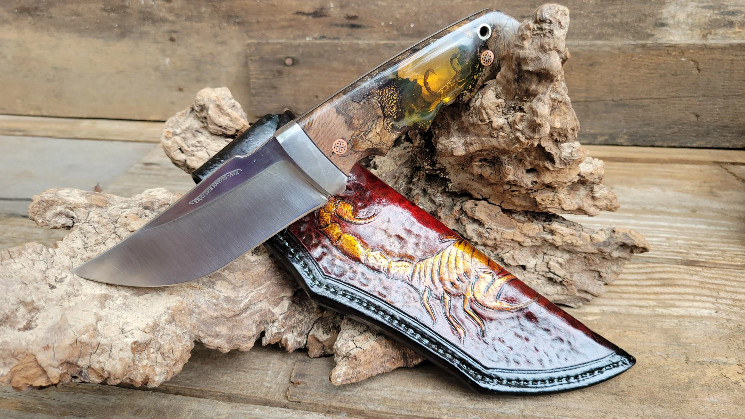 A fixed blade knife with a wood and resin handle resting on pieces of driftwood and a decorated sheath with a scorpion design on a wooden surface.