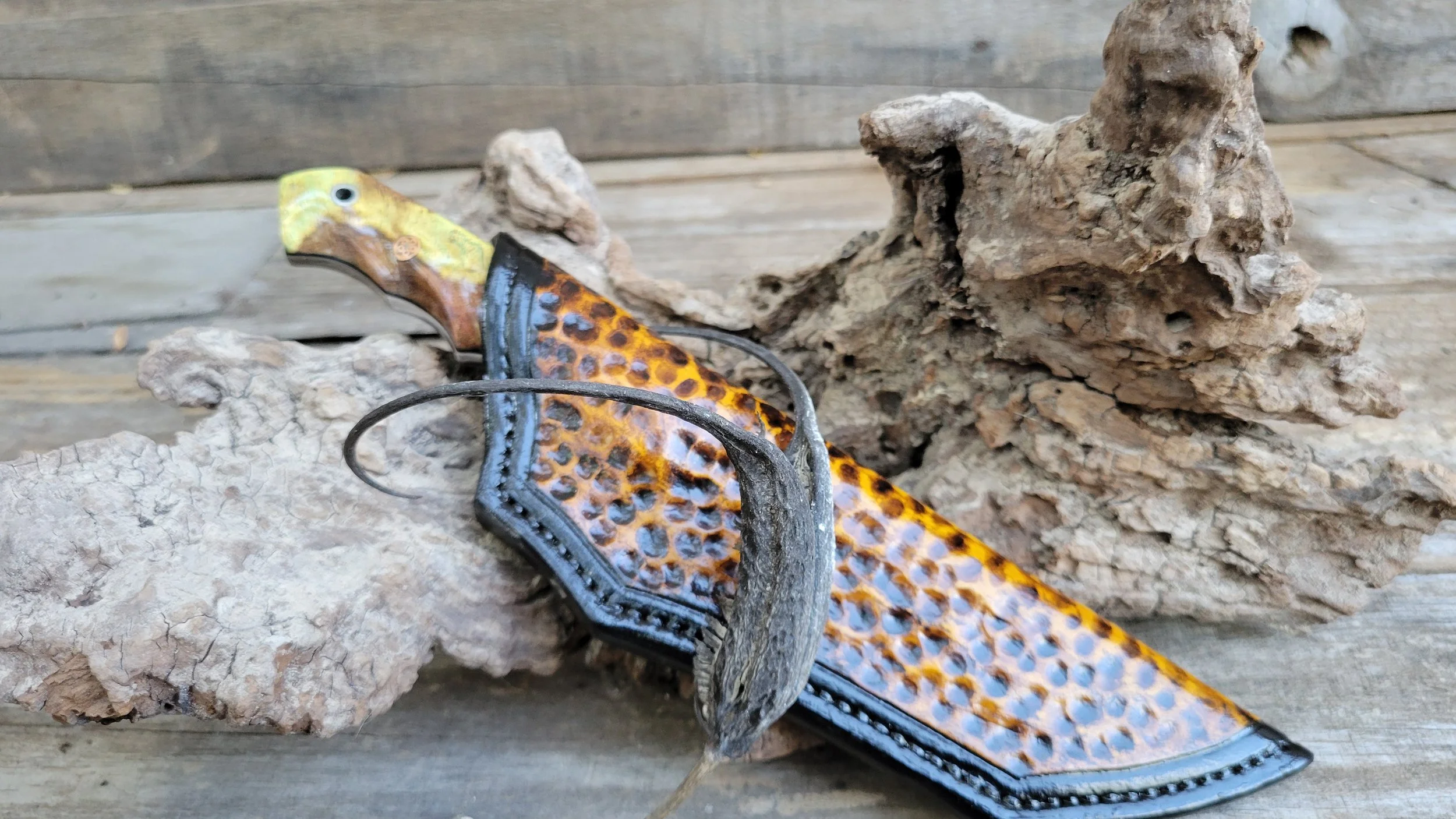 Decorative knife with a brown and yellow patterned blade, brass handle, and an attached strap, resting on textured rocks on a wooden surface.