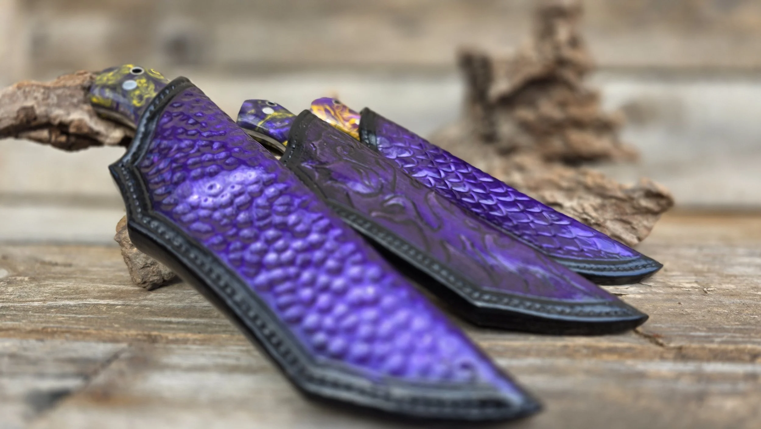 Three decorative knives with purple snake-skin pattern handles and black detailing, resting on a weathered wooden surface with dried leaves in the background.