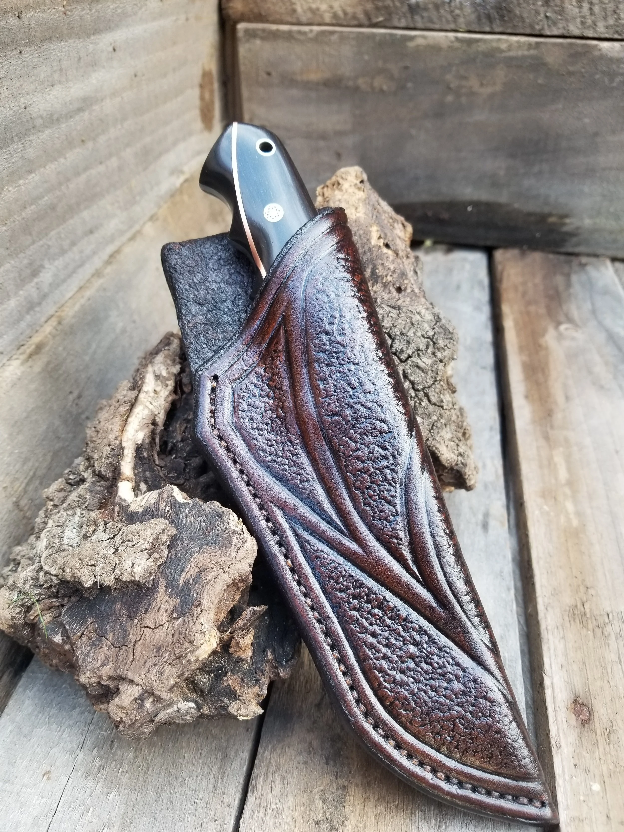 A folding knife partially tucked into a leather sheath shaped like a leaf, resting on a piece of bark on a wooden surface.