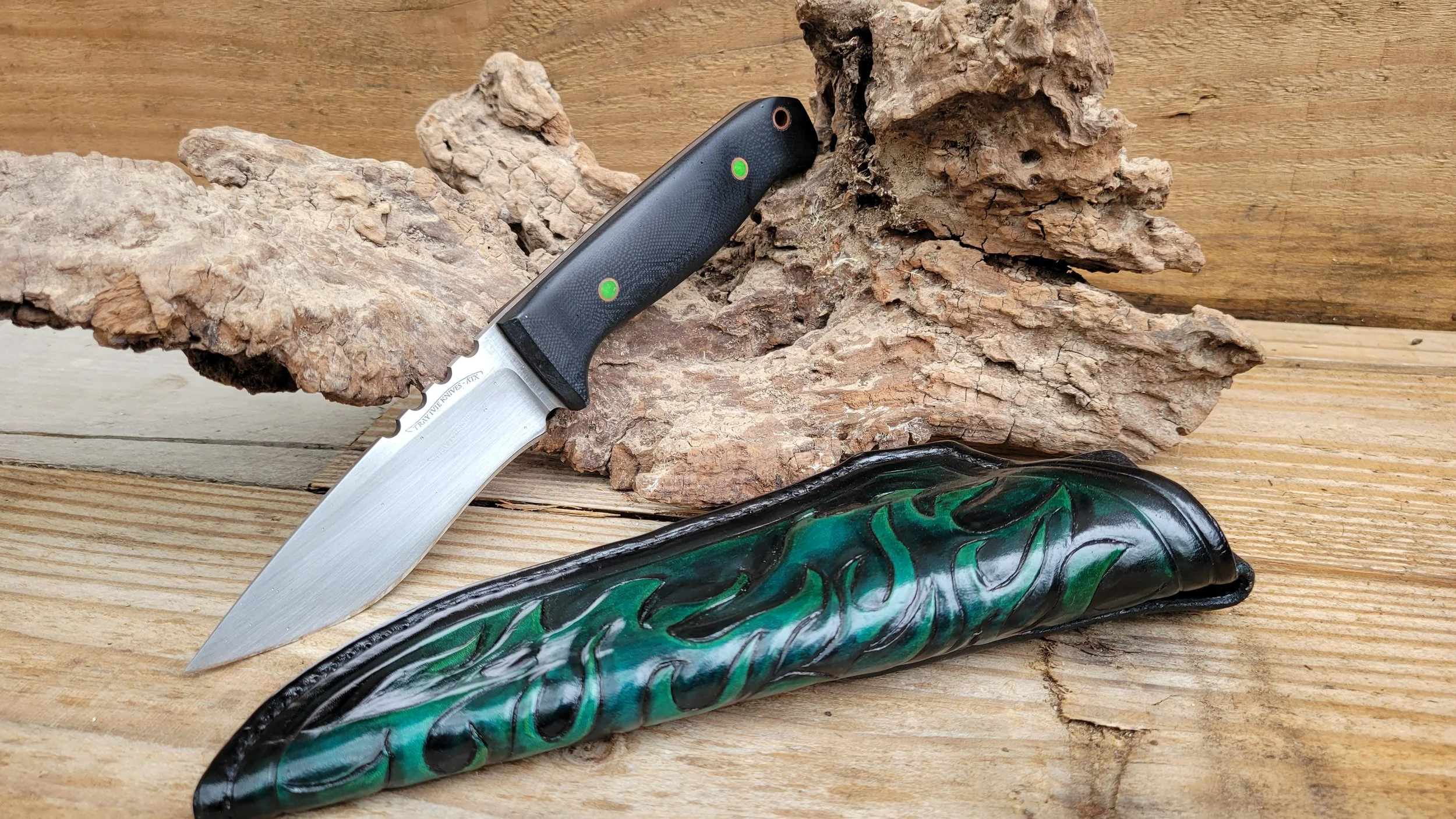 A hunting knife with a black handle and silver blade rests on a piece of driftwood, with a carved green and black shell case below it on a wooden surface.