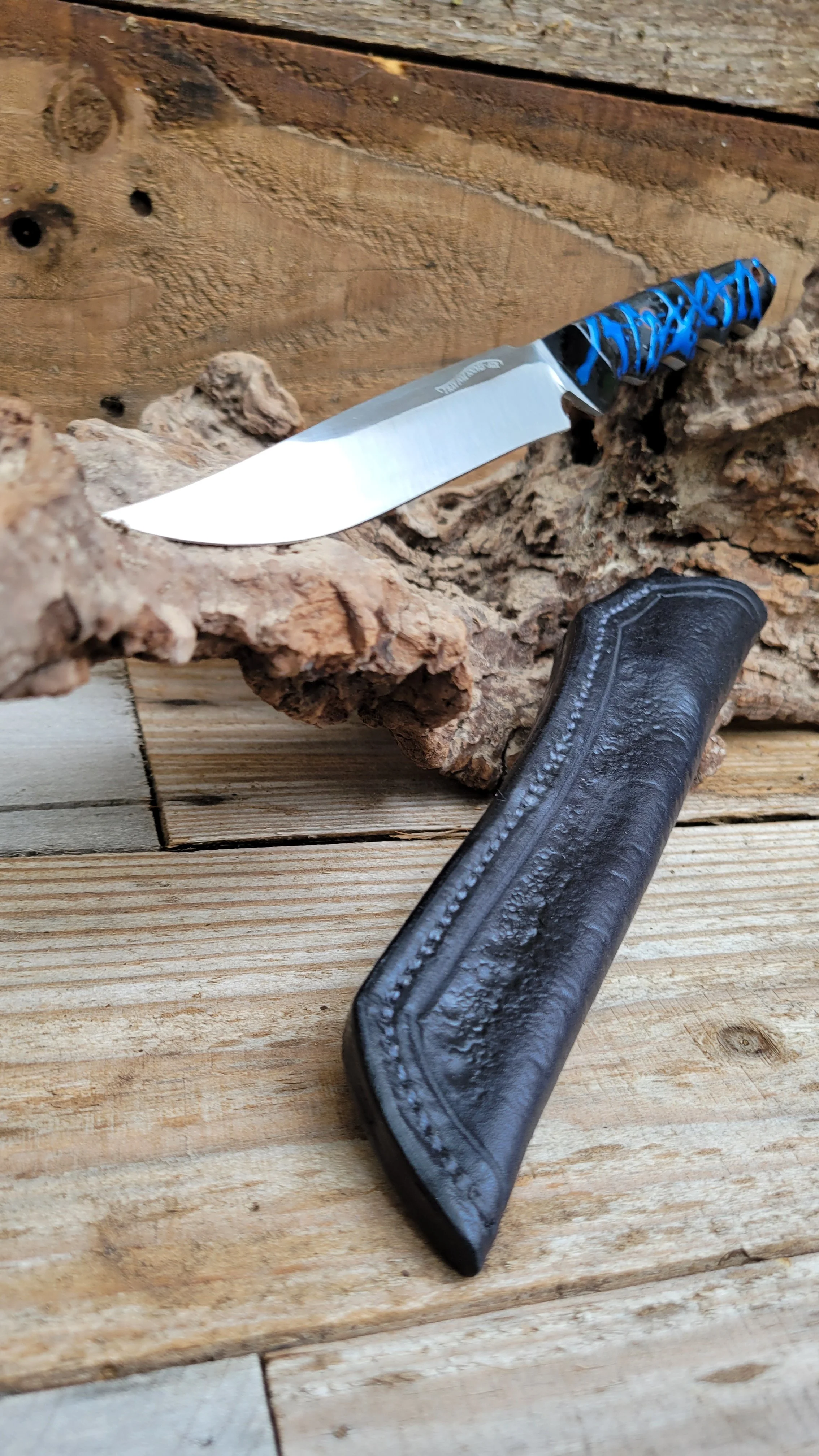 A fixed blade knife with a black and blue handle and a shiny silver blade, resting on a piece of rough bark, with a black leather sheath lying in front.