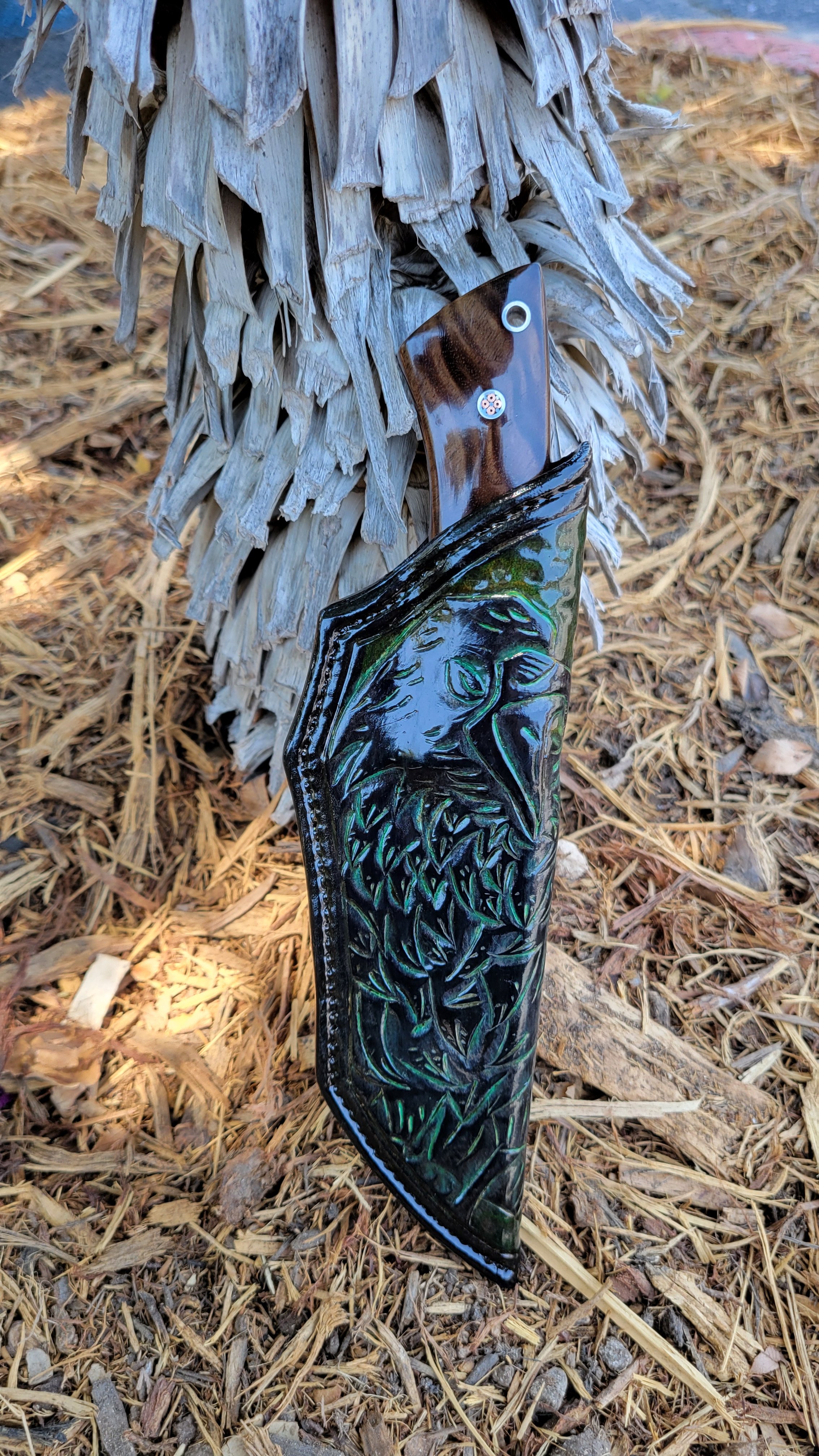 A decorative knife with a carved wooden handle and a molded snake design on the blade is leaning against a palm tree trunk in a dry, leaf-strewn outdoor area.