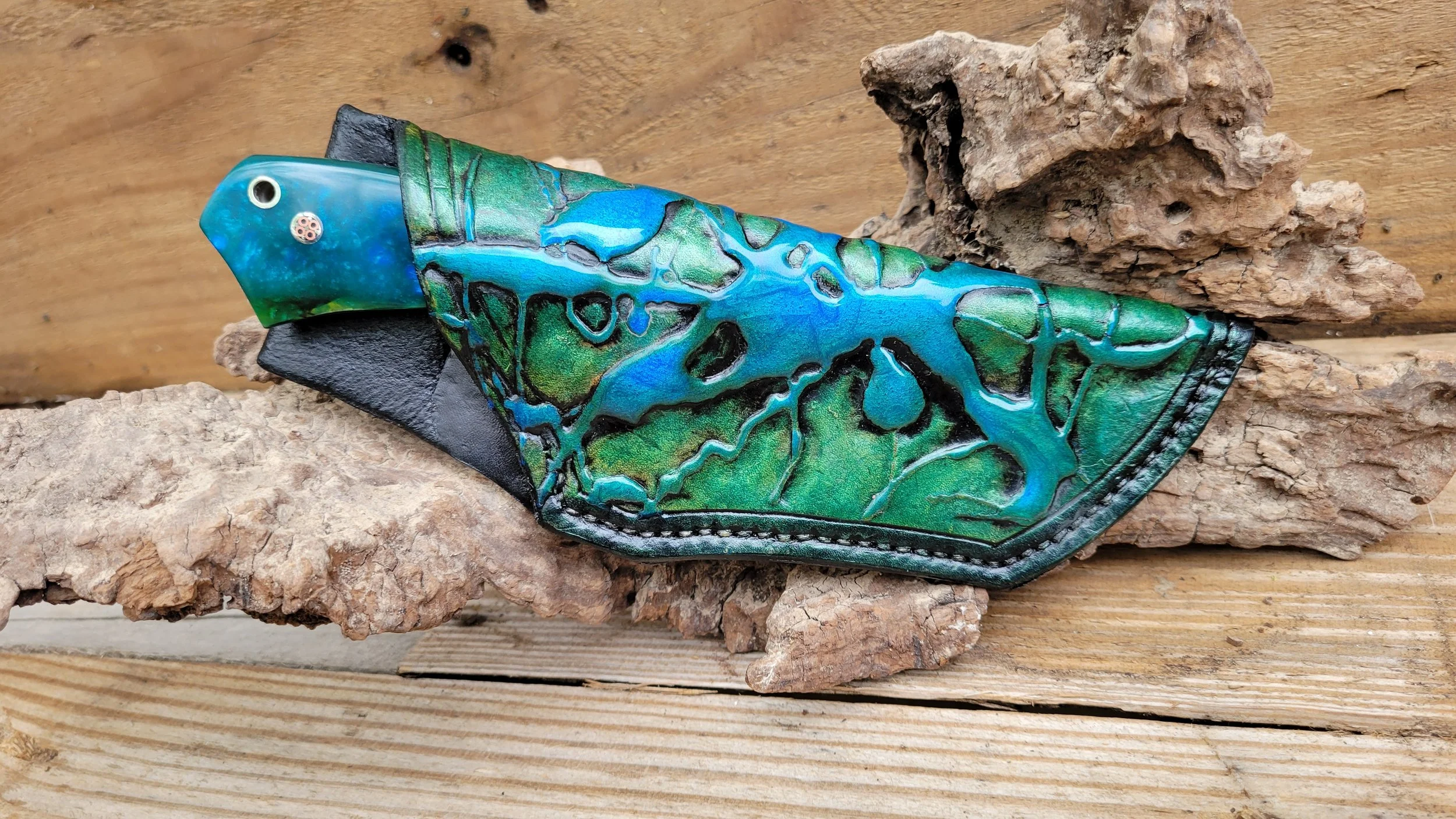 A knife with a handle made of blue and green marbled material and a blade covered with a detailed, colorful, abstract design resembling a landscape, resting on rocks and a wooden surface.