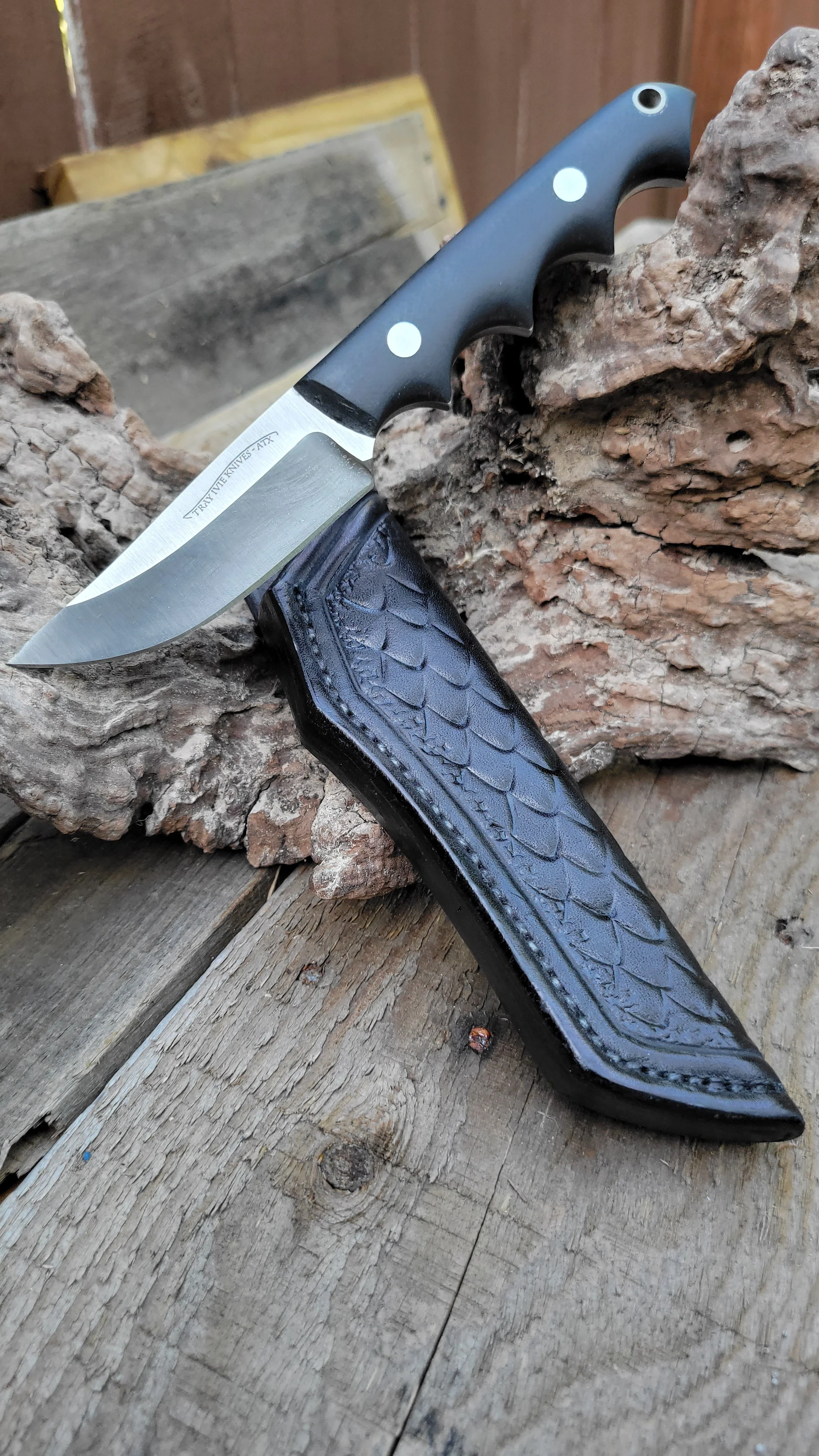 A fixed-blade knife with a black handle resting on a log, with a leather sheath placed underneath.