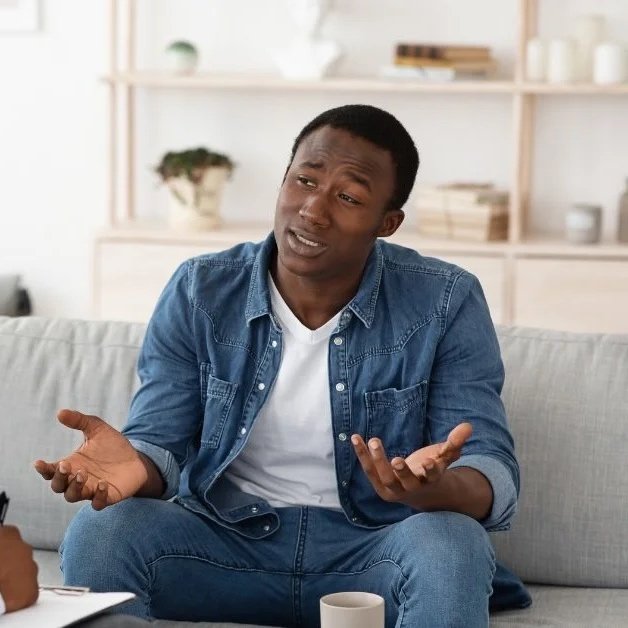 A man sitting on a gray couch, wearing a denim shirt and white t-shirt, gesturing with his hands while talking, in a bright living room with shelves and decorative items in the background.