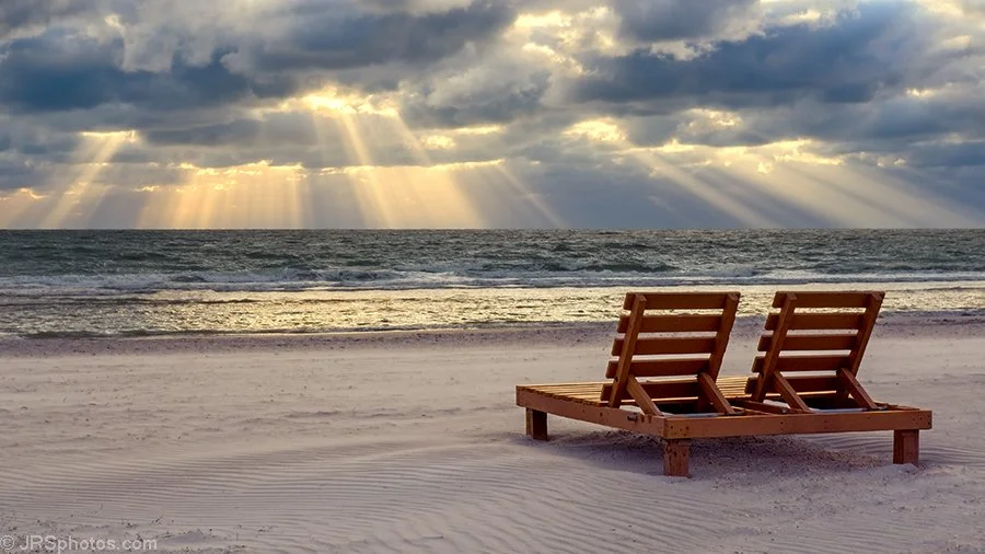 Two wooden lounge chairs on a sandy beach facing the ocean under a cloudy sky with sun rays