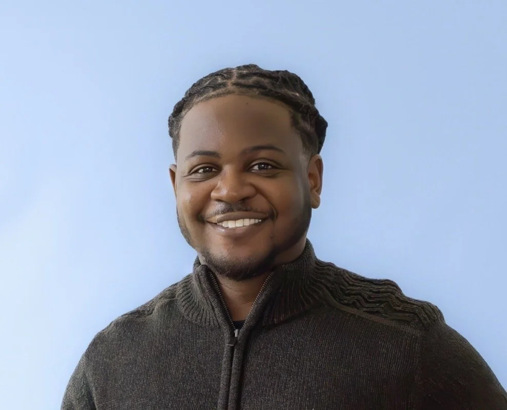 Smiling man with braided hair wearing a black zip-up sweater against a light blue background.