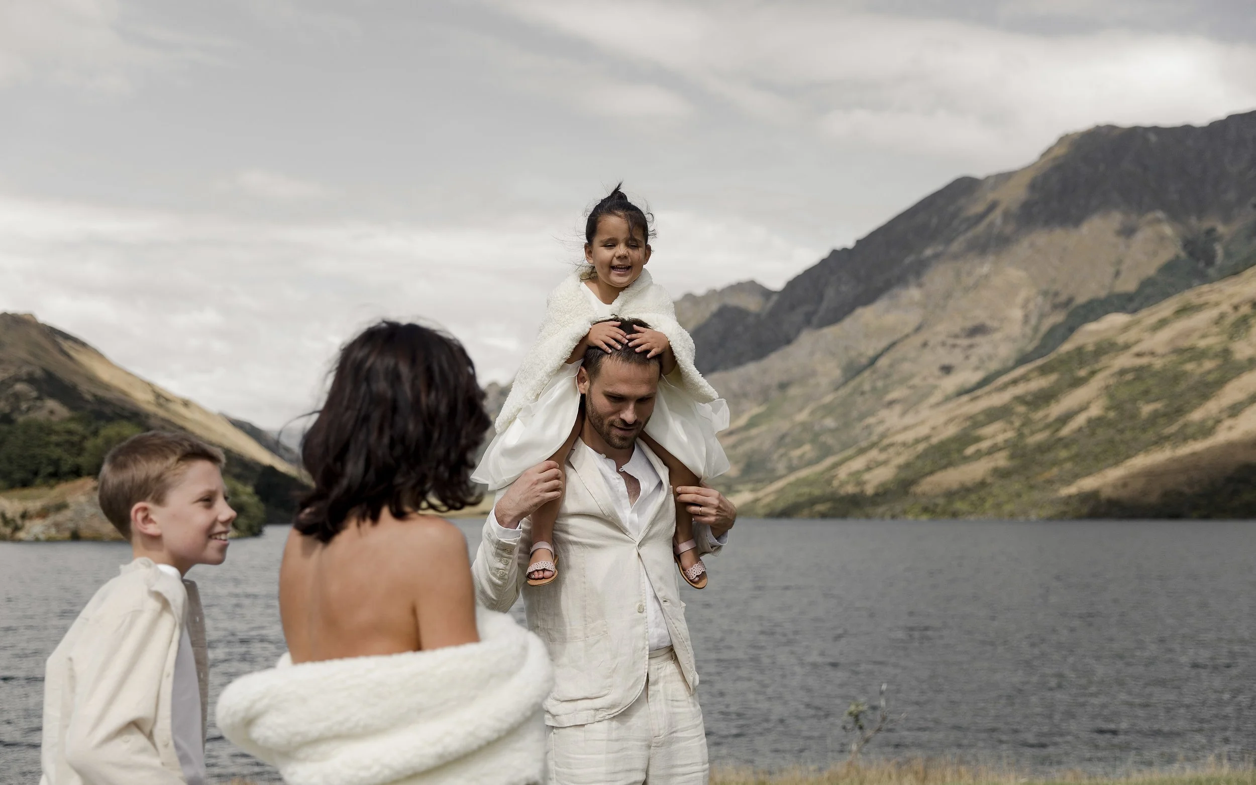 A man carrying a young girl on his shoulders near a lake with mountains in the background, with a woman and a young boy nearby, all outdoors in a scenic mountainous area.