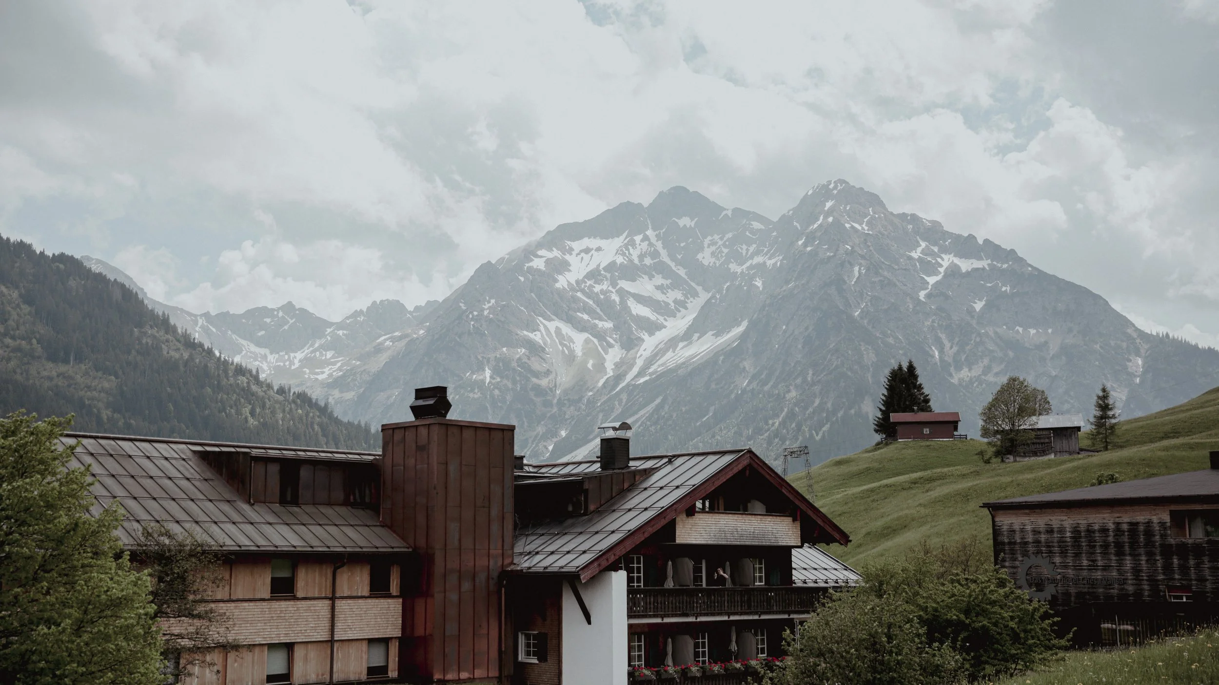Mountain landscape with snow-capped peaks, green hills, and a few houses at the foreground.