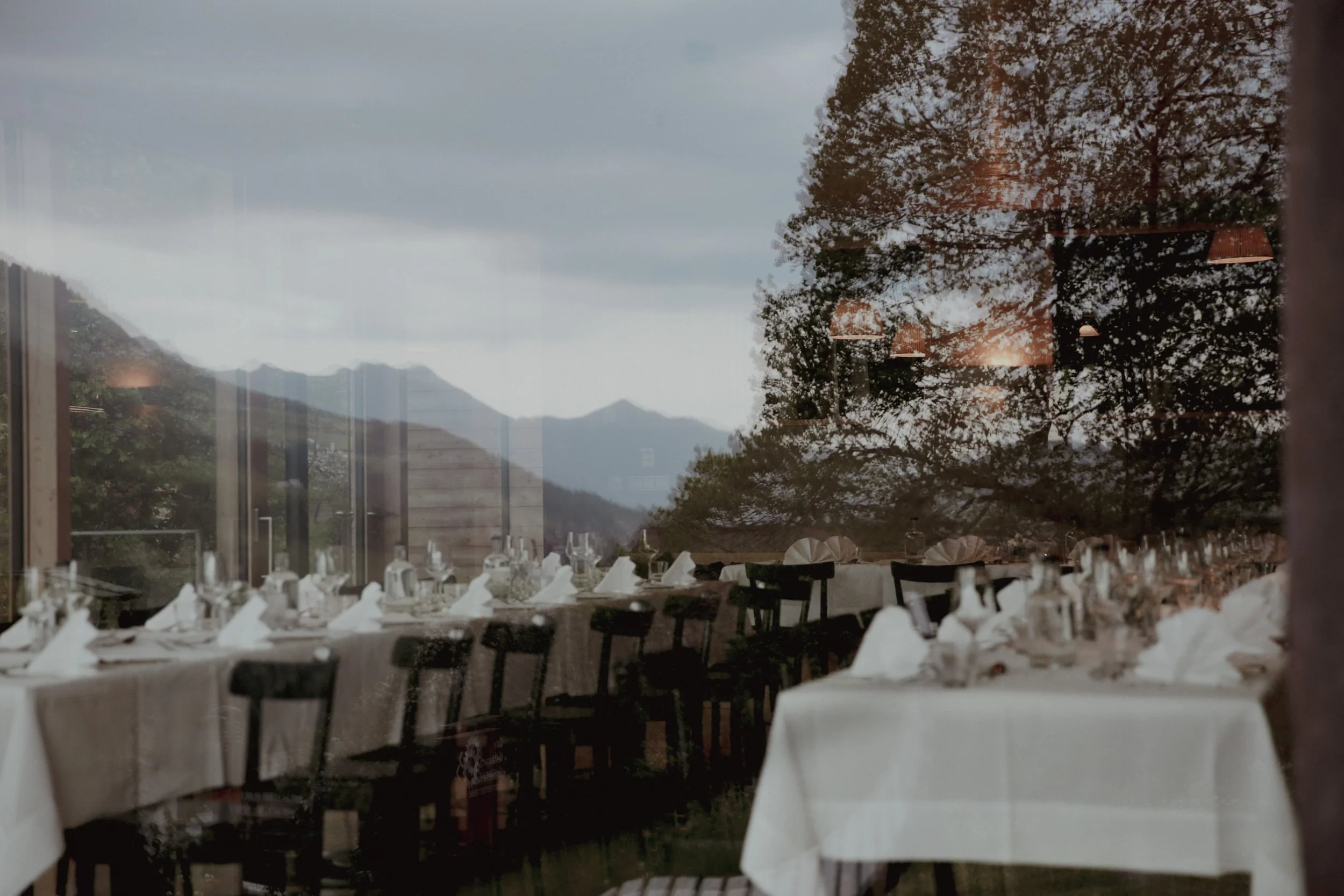 Empty restaurant with white-draped tables, glassware, and napkins, reflected in a window showing a mountainous landscape and a large tree with leaves in autumn colors.