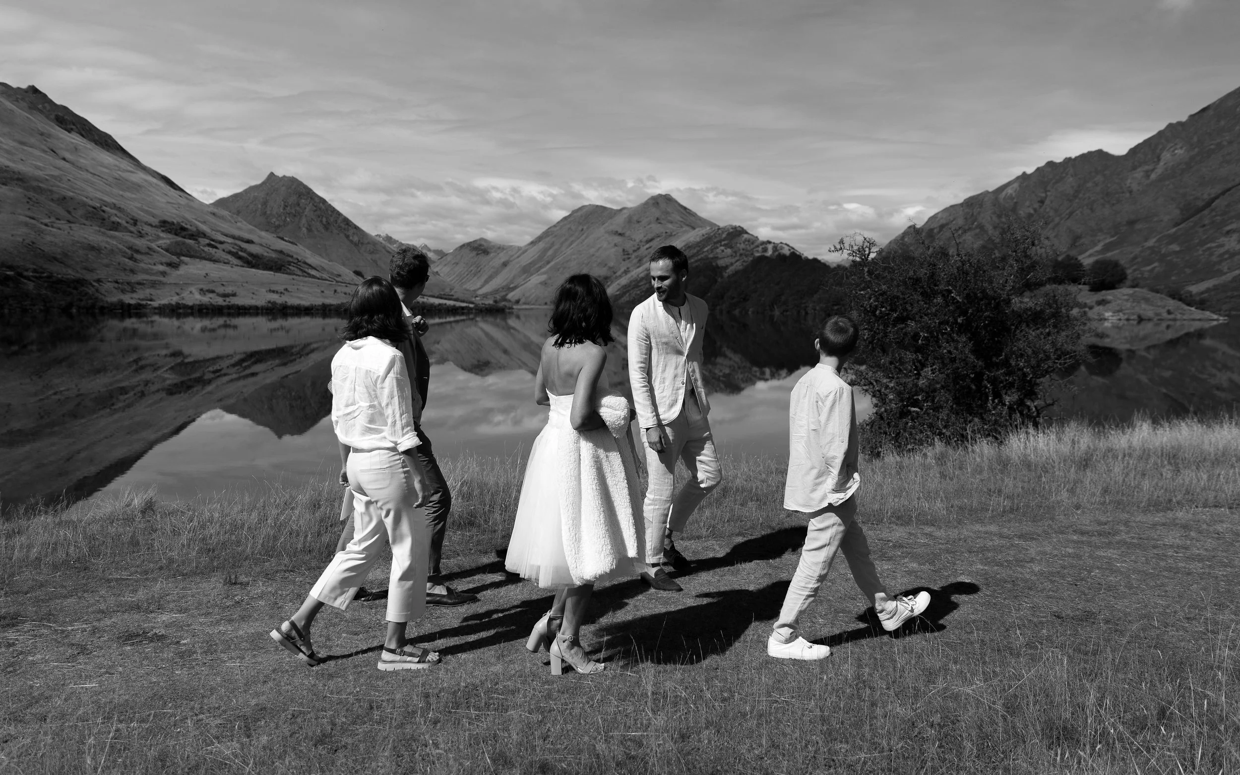Black and white photo of a wedding party walking along a grassy area by a lake with mountains in the background. The bride is in a white dress, and the groom and others are dressed in formal attire.
