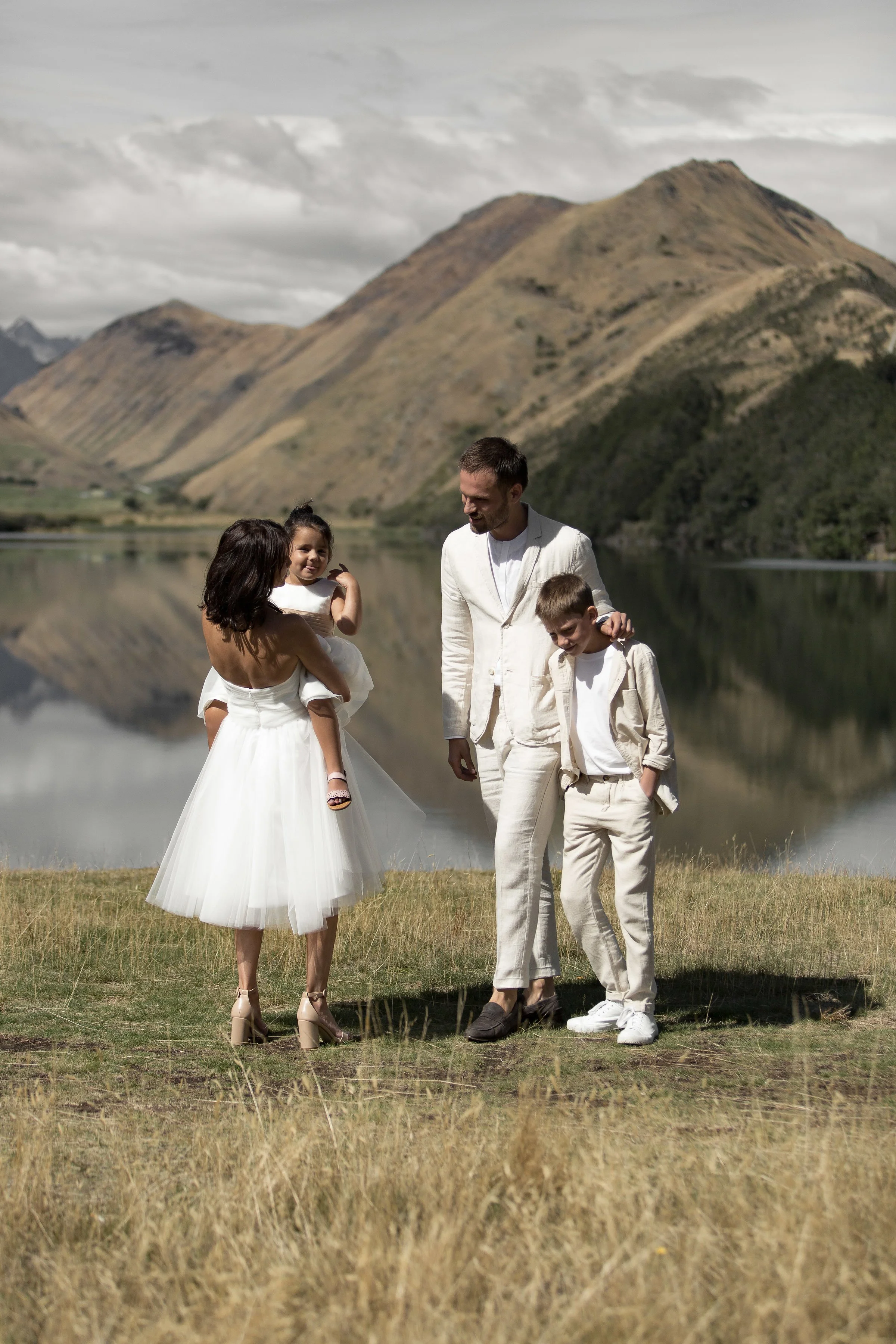 A family of four standing on grass near a lake with mountains in the background. The mother holds a young girl, and the father stands next to an older boy. They are dressed in light-colored clothing.