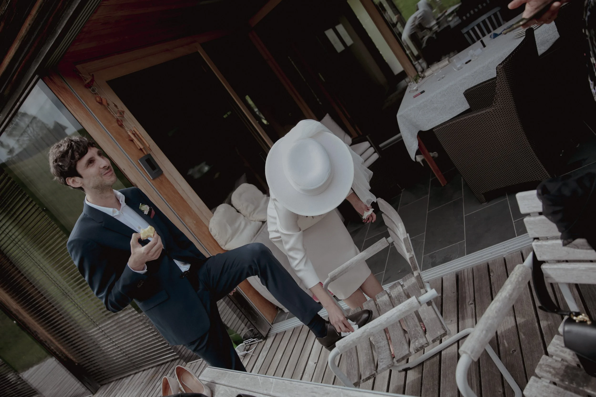 A man in a navy blue suit holding a piece of food and a woman in a white dress and hat reaching to adjust her shoe on a wooden deck.