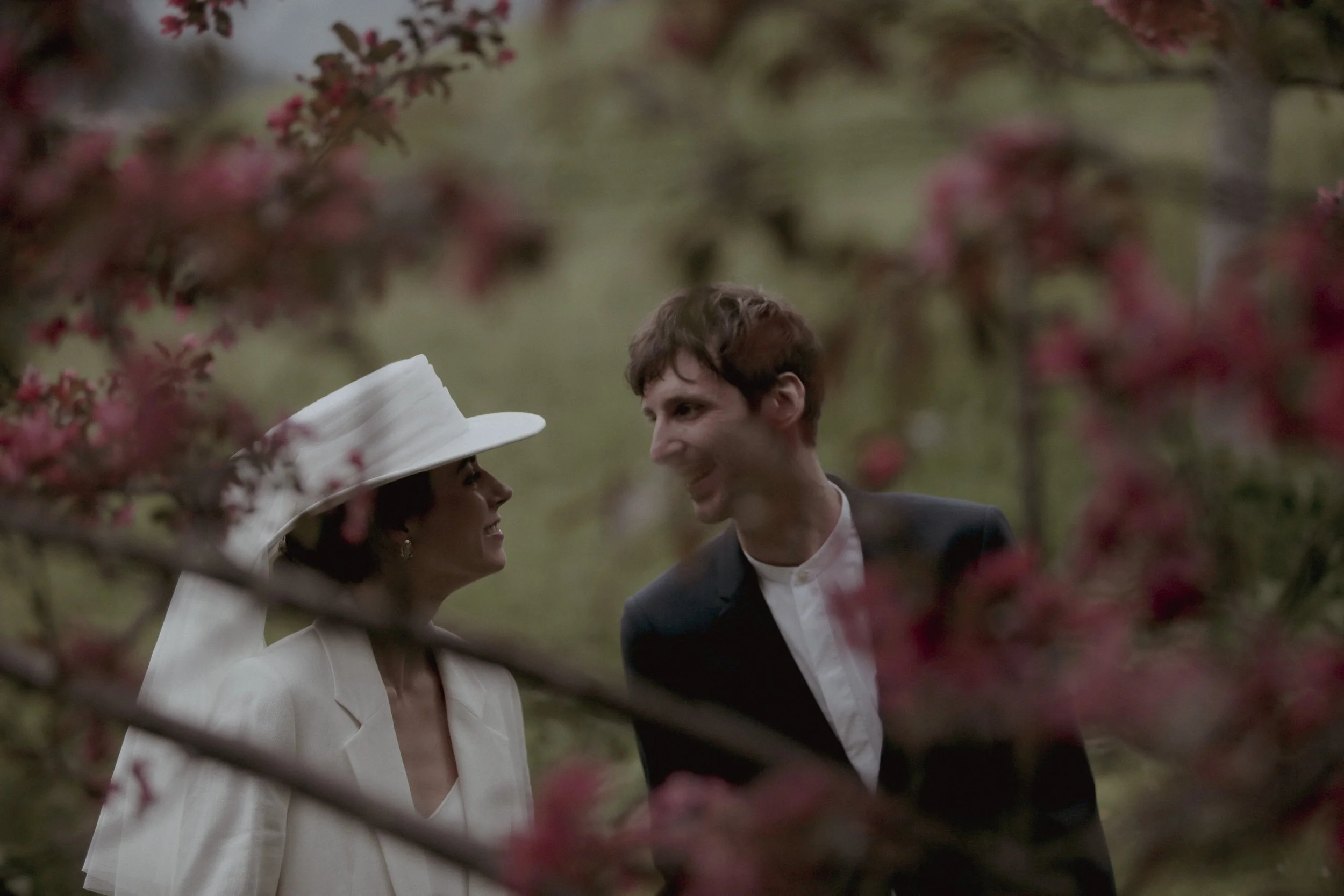 A woman in a white outfit and large white hat, and a man in a black blazer with a white shirt, smiling at each other among pink flowering trees.