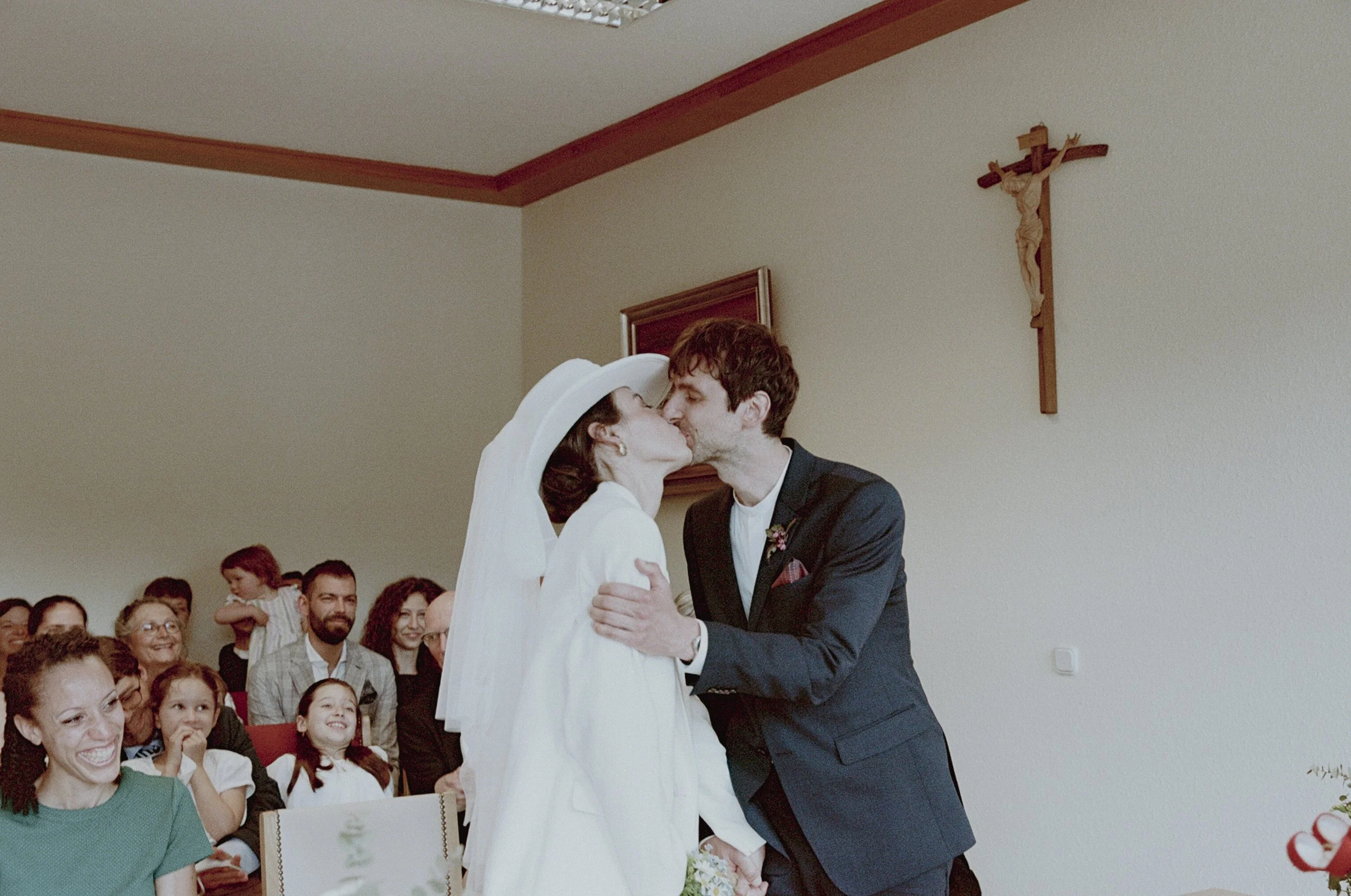 A couple in wedding attire share a kiss during their wedding ceremony in a church with an audience in the background.
