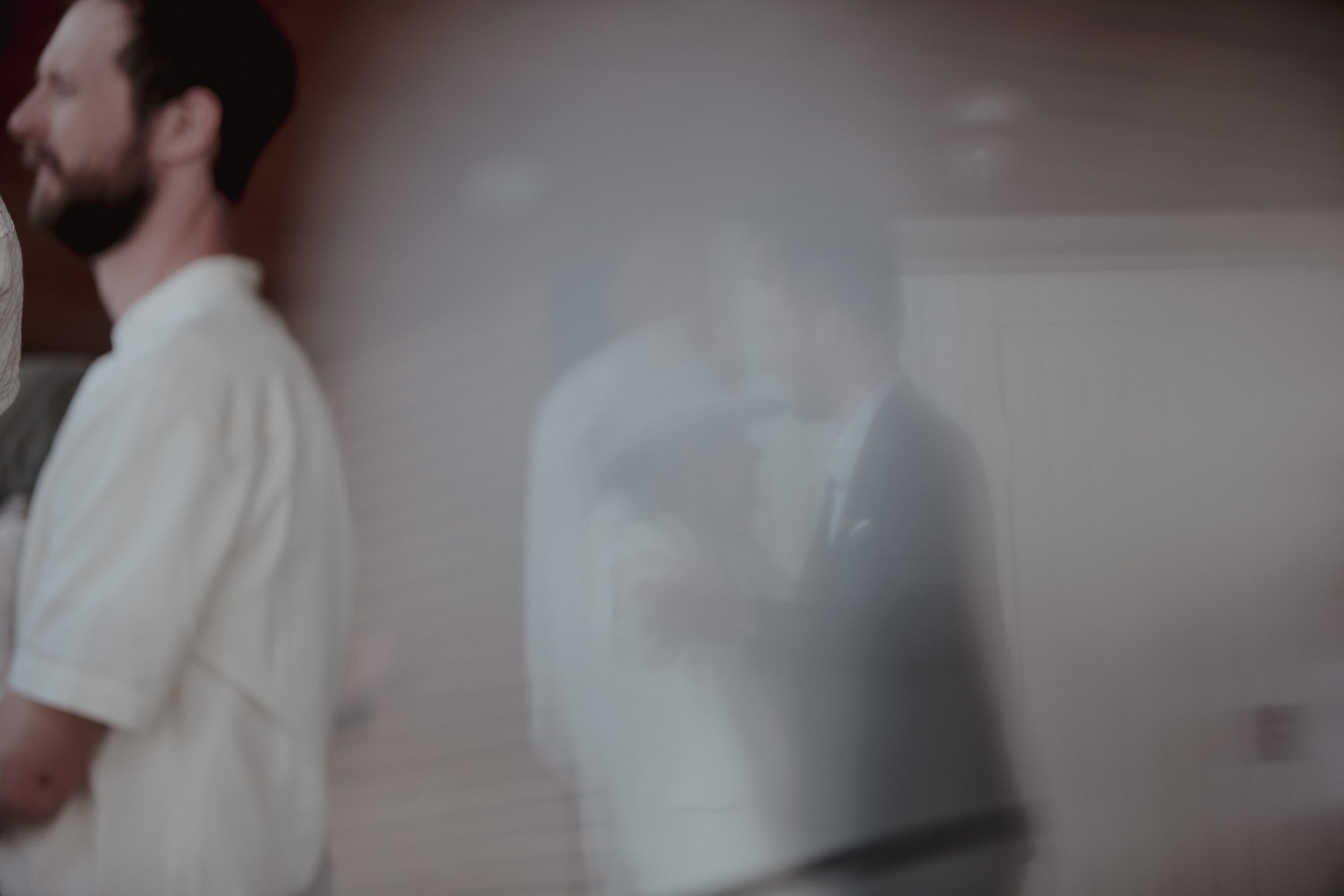 A man with a beard and white shirt standing against a wall, with his reflection appearing on a shiny surface in front of him.