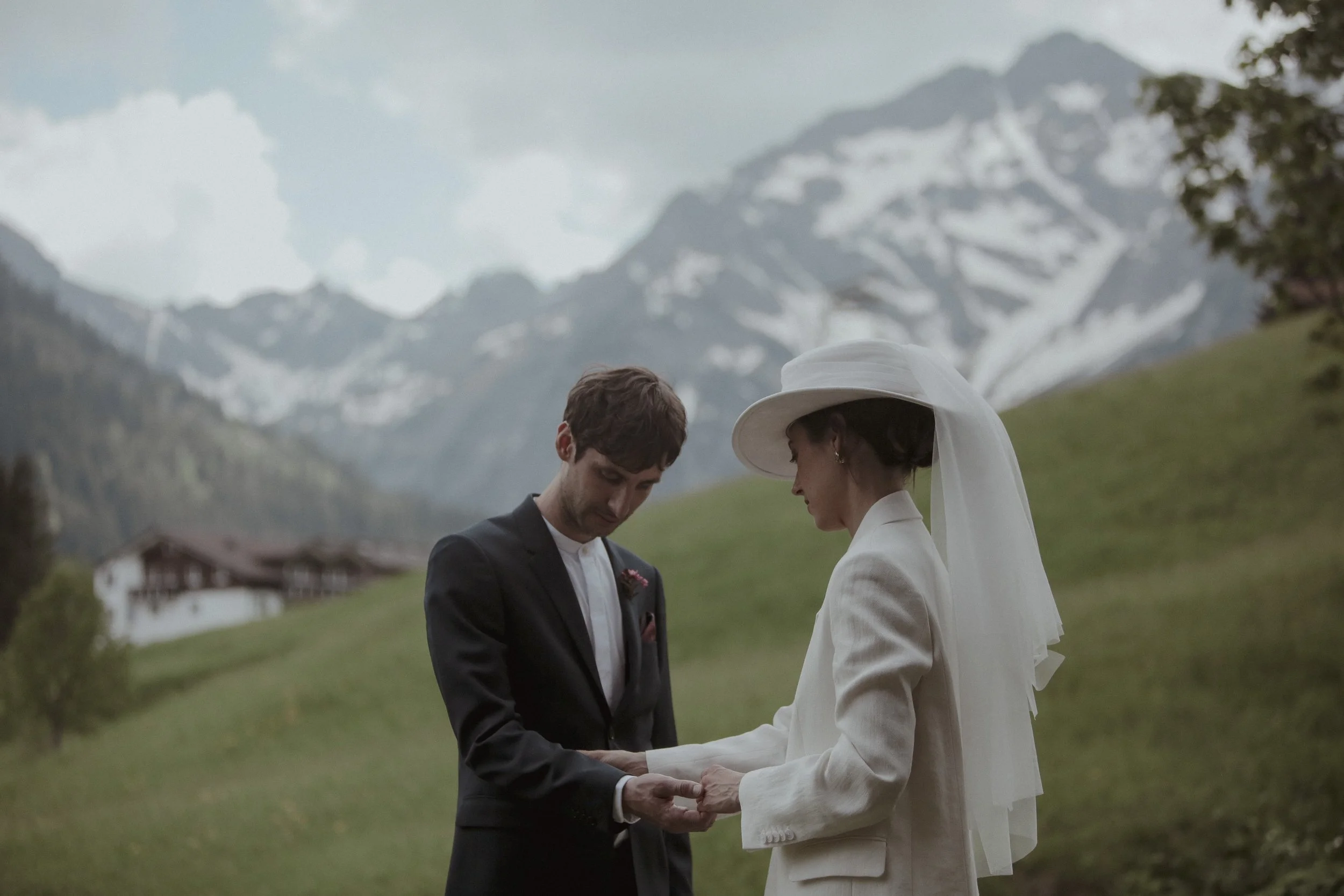 A bride and groom stand outdoors in a mountainous landscape during their wedding ceremony. The groom wears a dark suit, and the bride wears a white suit and large hat with a veil. They hold hands and look at each other.