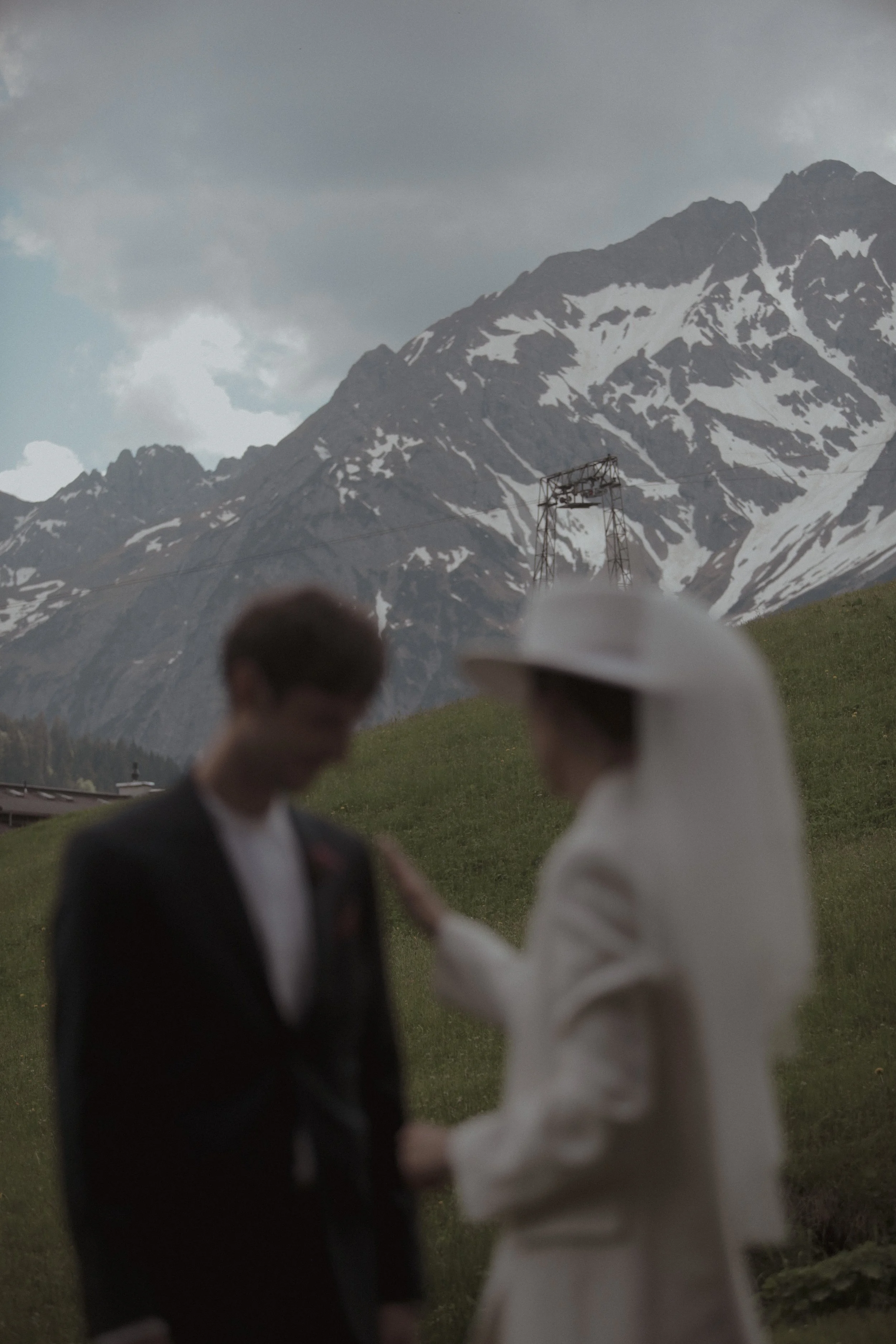 Two people, possibly a couple, standing outdoors with mountains and snow in the background. One person is wearing a white hat and coat, the other a dark suit. Their faces are blurred, creating a romantic or intimate scene.