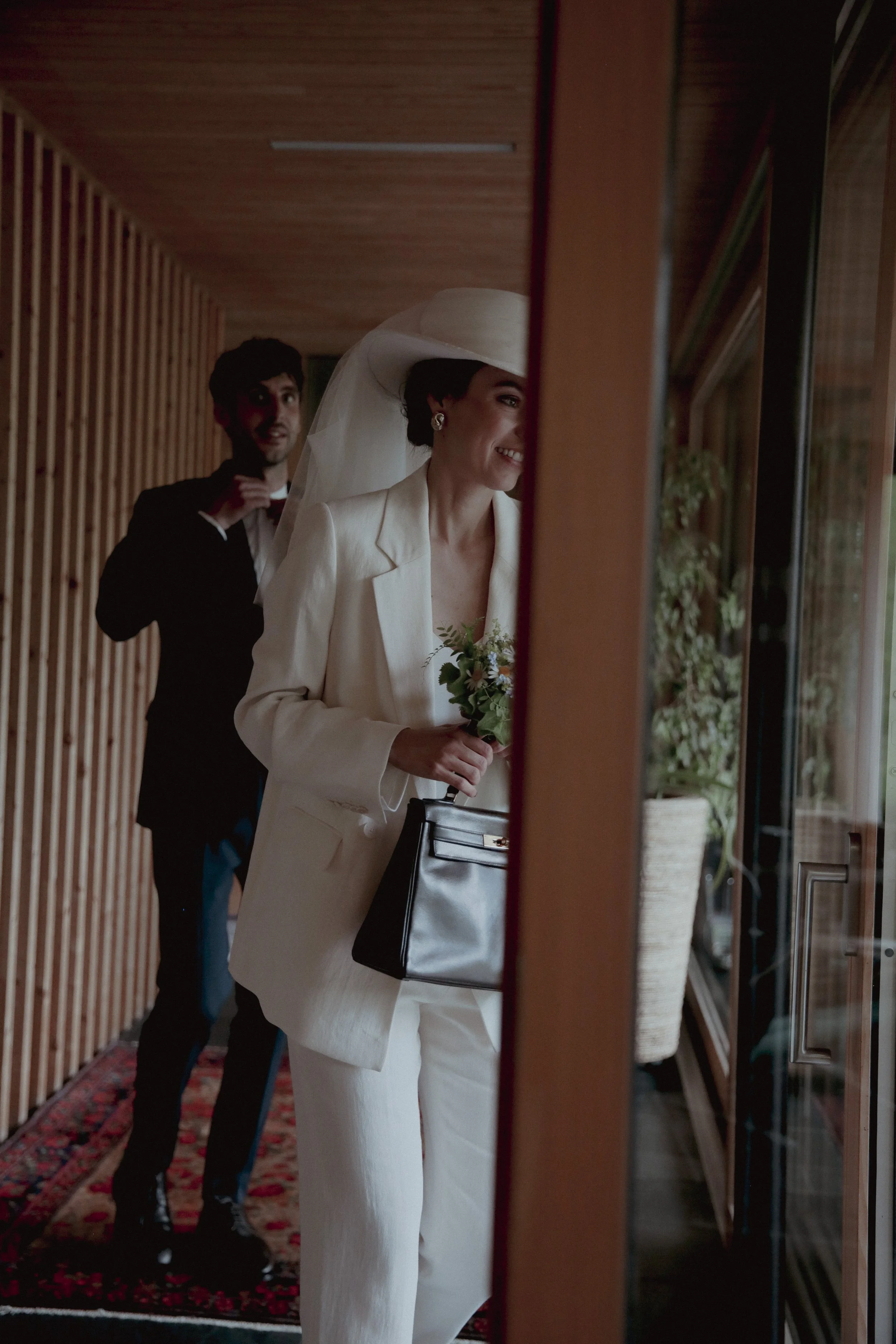 A bride in a white suit with a large hat and veil, holding a bouquet, is smiling and looking out a window, with a man in a suit adjusting his tie behind her.