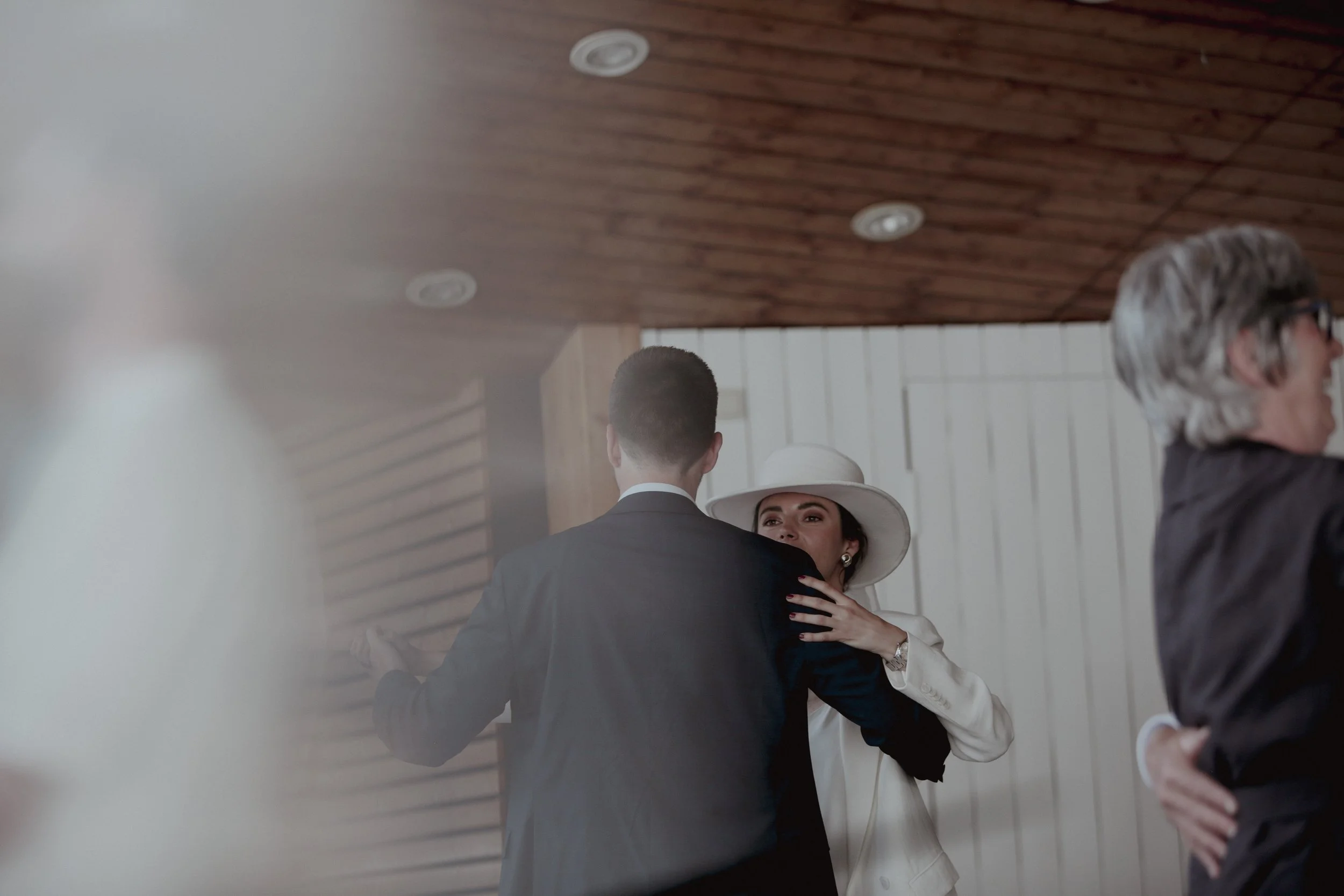 A woman wearing a white hat and white blazer dancing with a man in a black suit. Other guests are visible in the background, gathered at a social event.