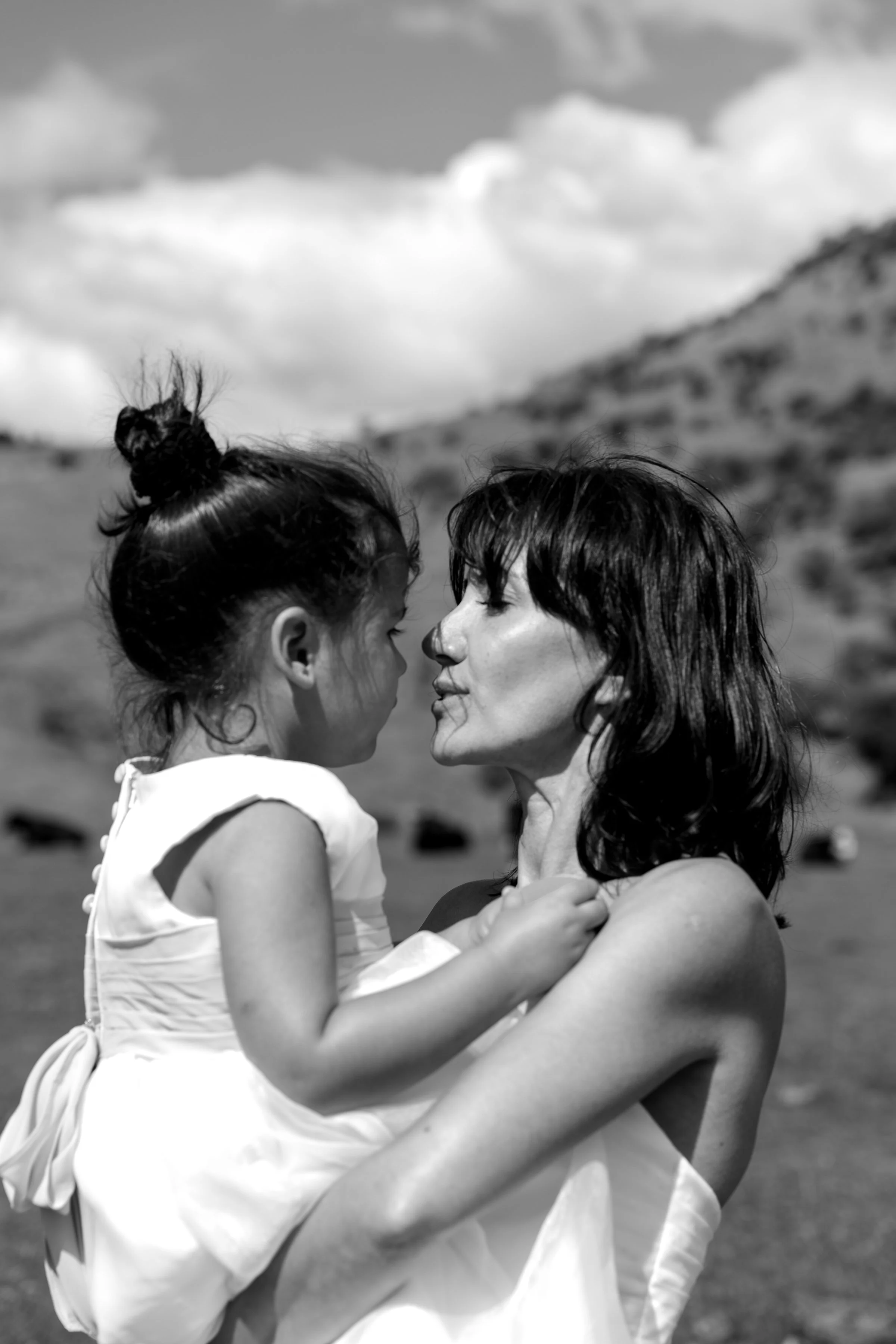 A black and white photo of a woman holding a young girl close, with their noses almost touching, outdoors with hills and cloudy sky in the background.