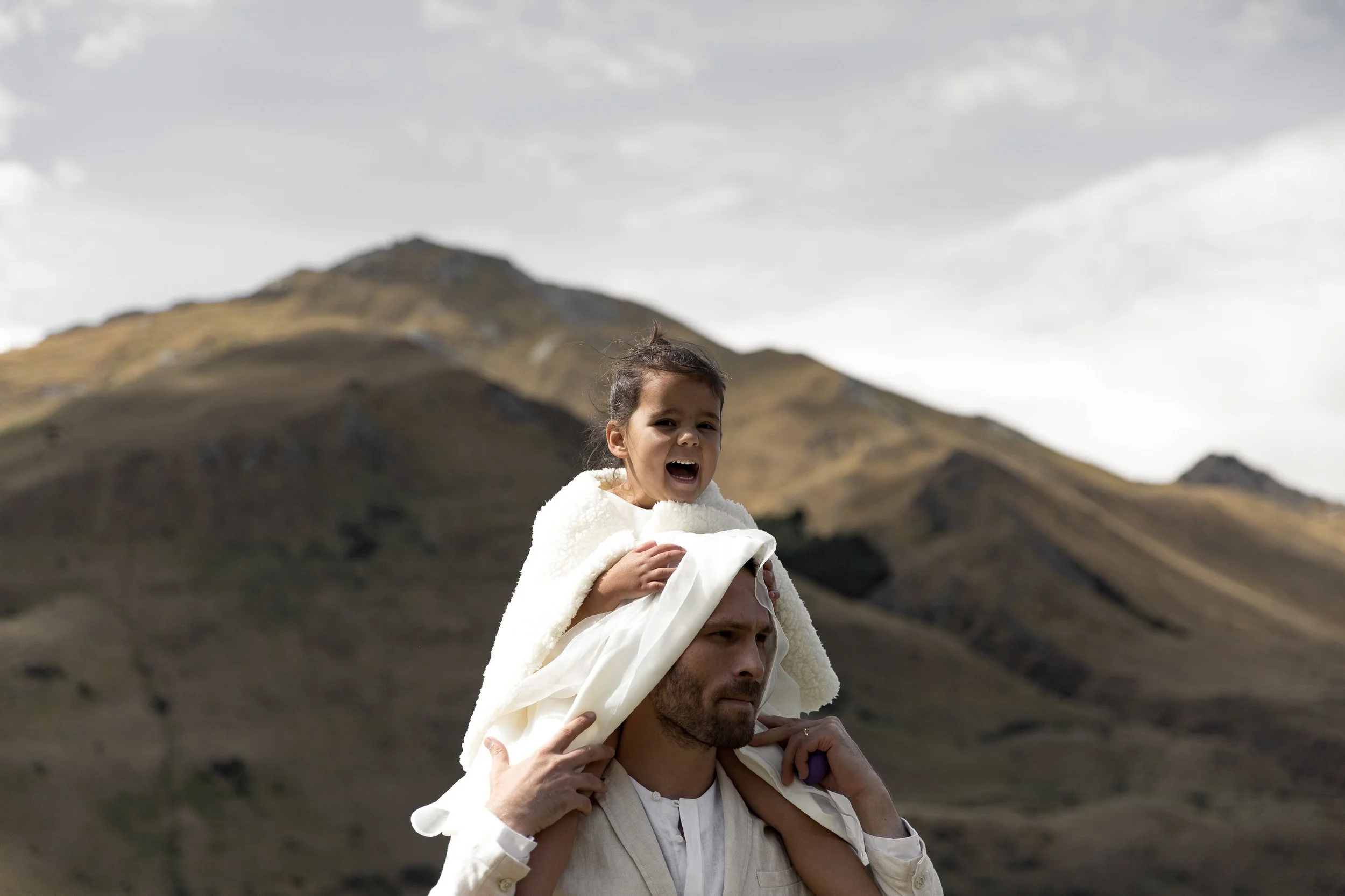 A man carrying a young girl on his shoulders outdoors with mountains in the background.