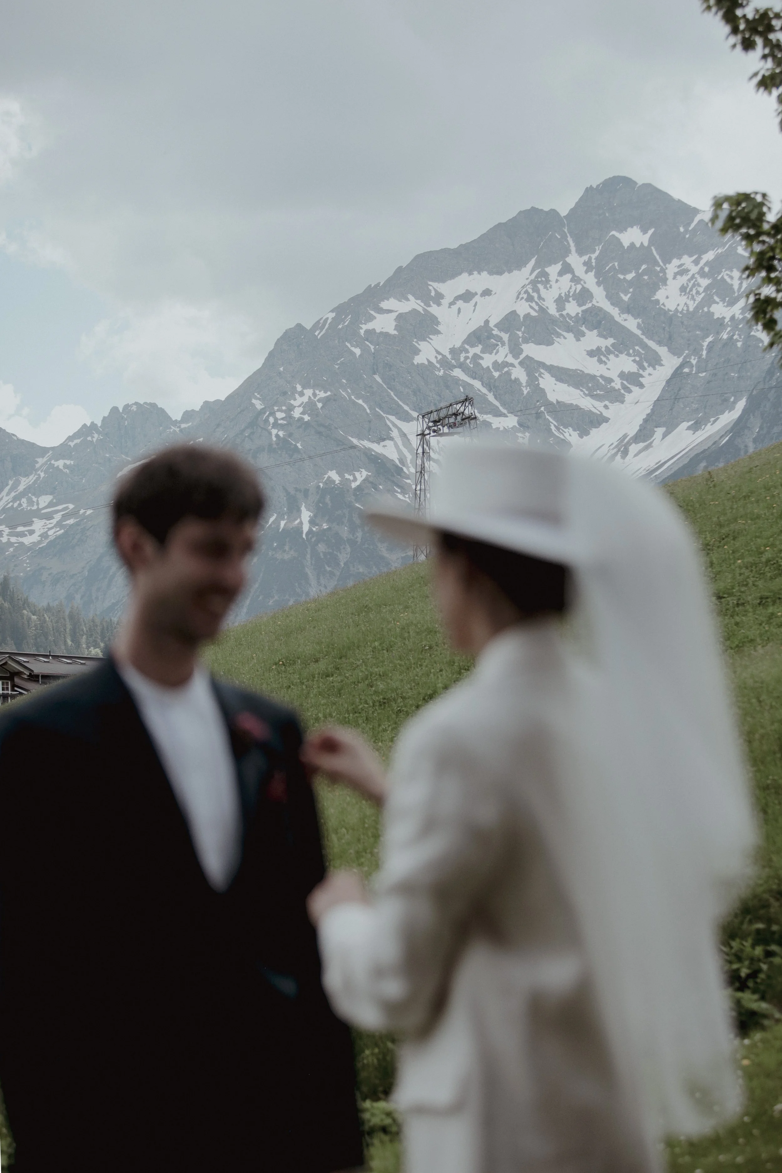 A blurry image of a couple, a man and a woman, standing outdoors on a grassy hillside with snow-capped mountains in the background, possibly during a wedding or special event.