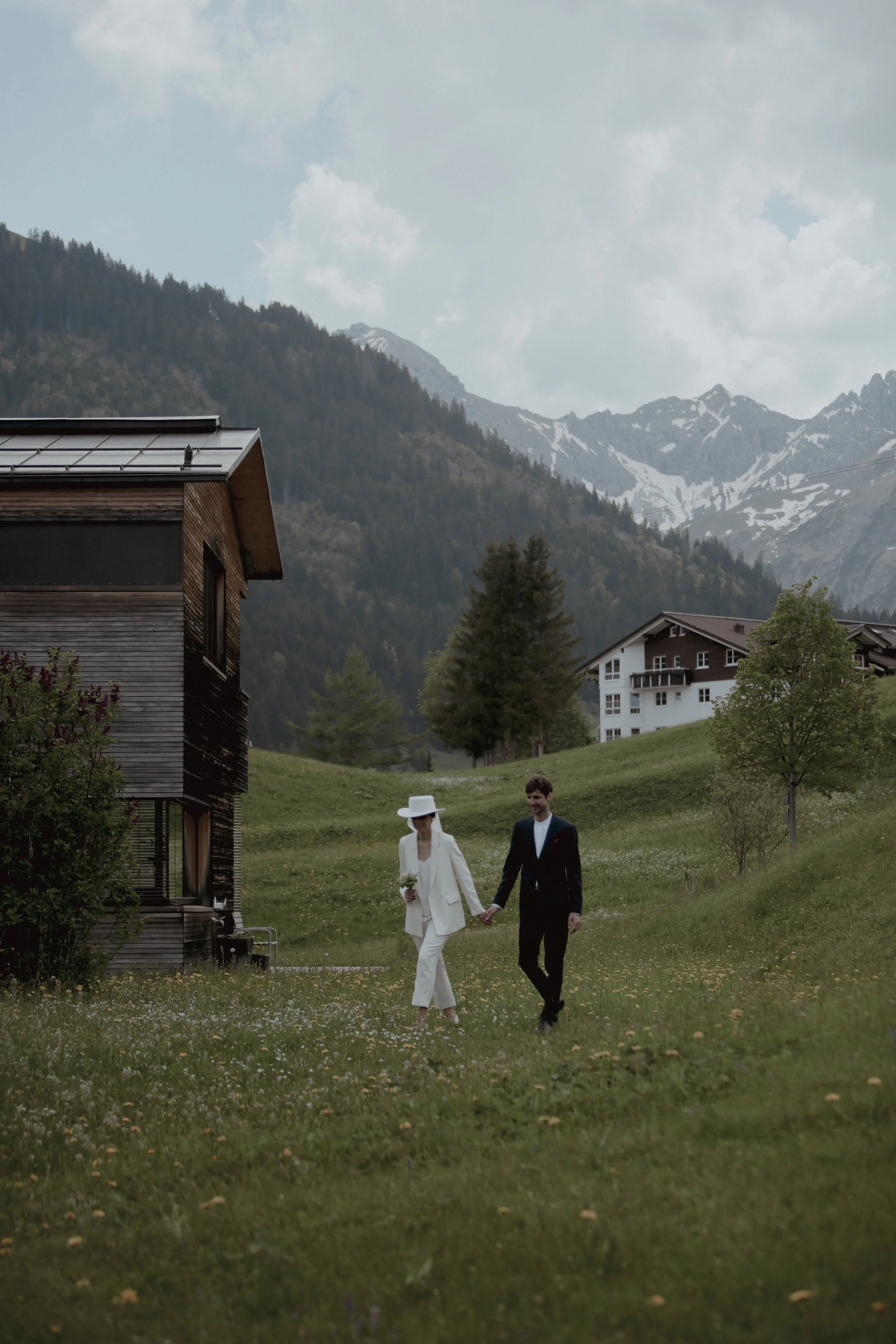 A bride and groom walking hand-in-hand through a grassy field with houses, trees, and mountains in the background.