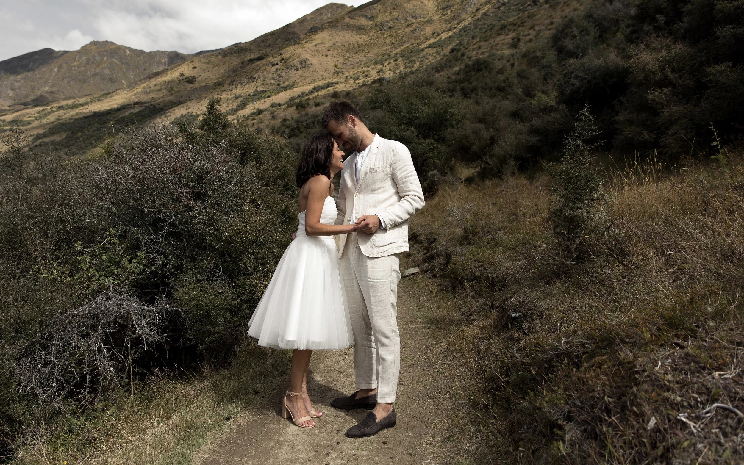 A couple in wedding attire standing close on a dirt trail in a mountainous area with dry grass and bushes.