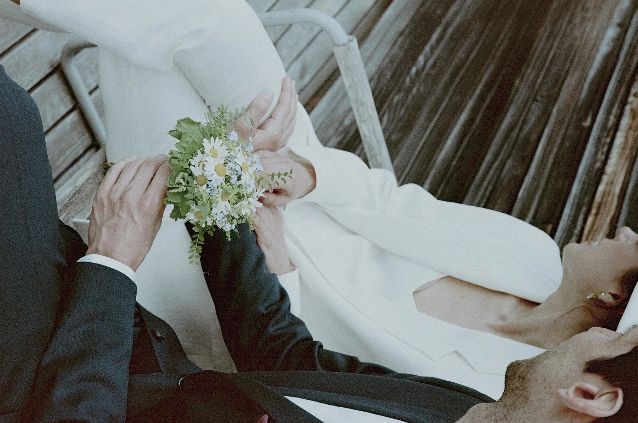A wedding scene with a bride in a white dress lying on a wooden deck, holding a bouquet of daisies and greenery, while a man in a dark suit and woman in black escort her.