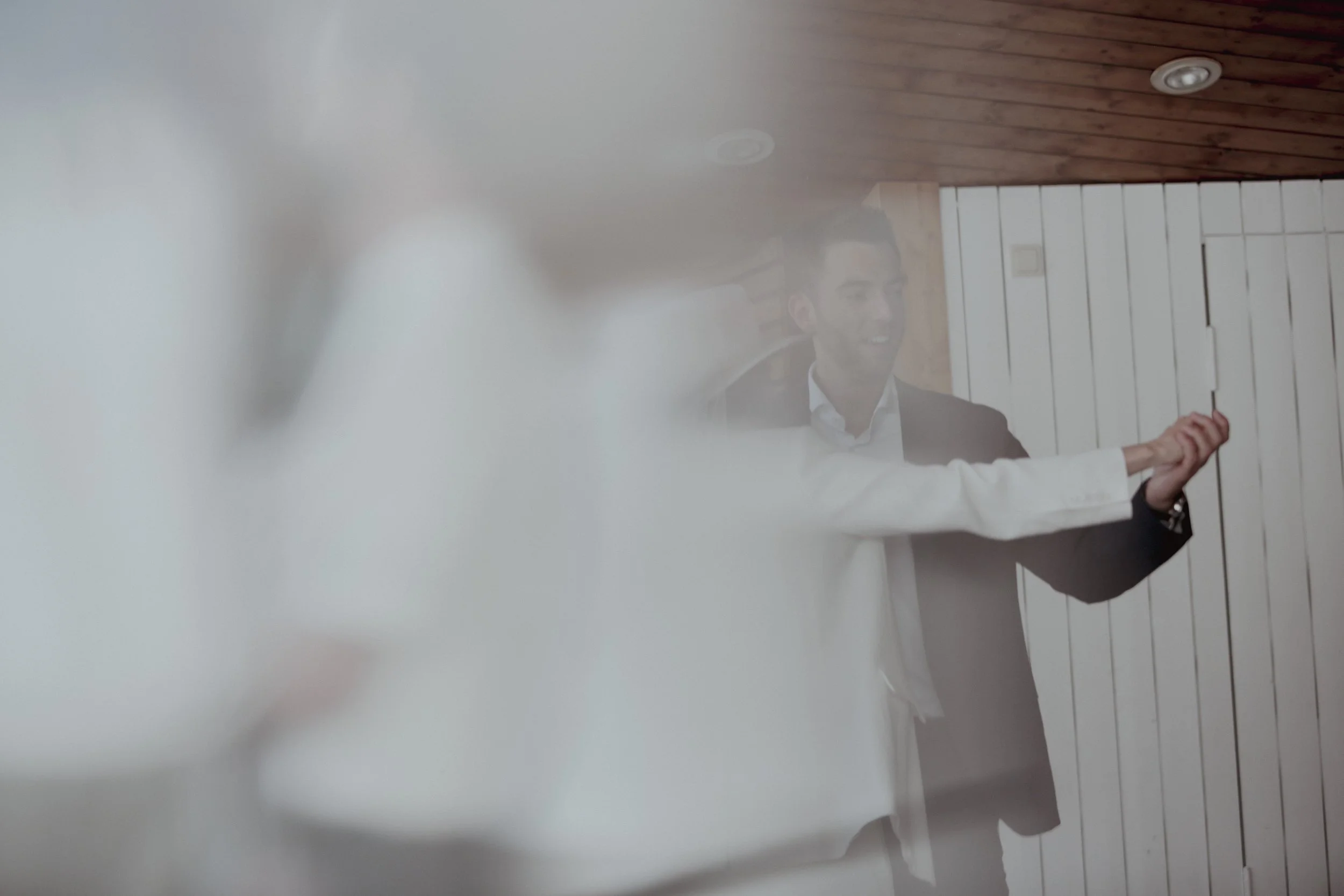 Young man in a suit smiling and extending his arm, seen through a blurred white flower or object.