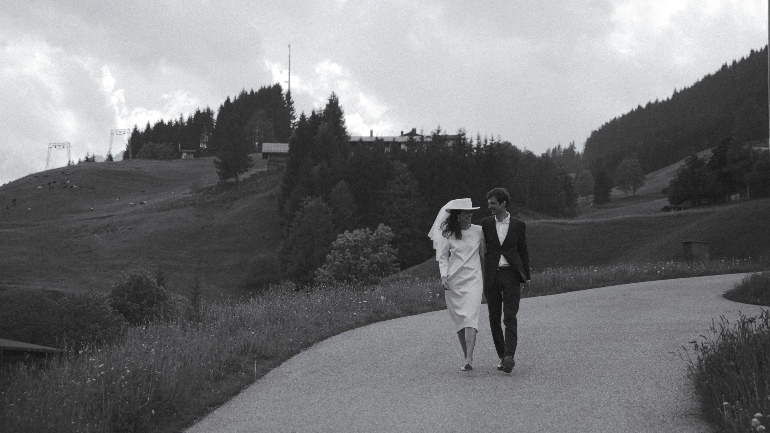 A black and white photo of a bride and groom walking together on a curved rural road with hills, trees, and houses in the background.
