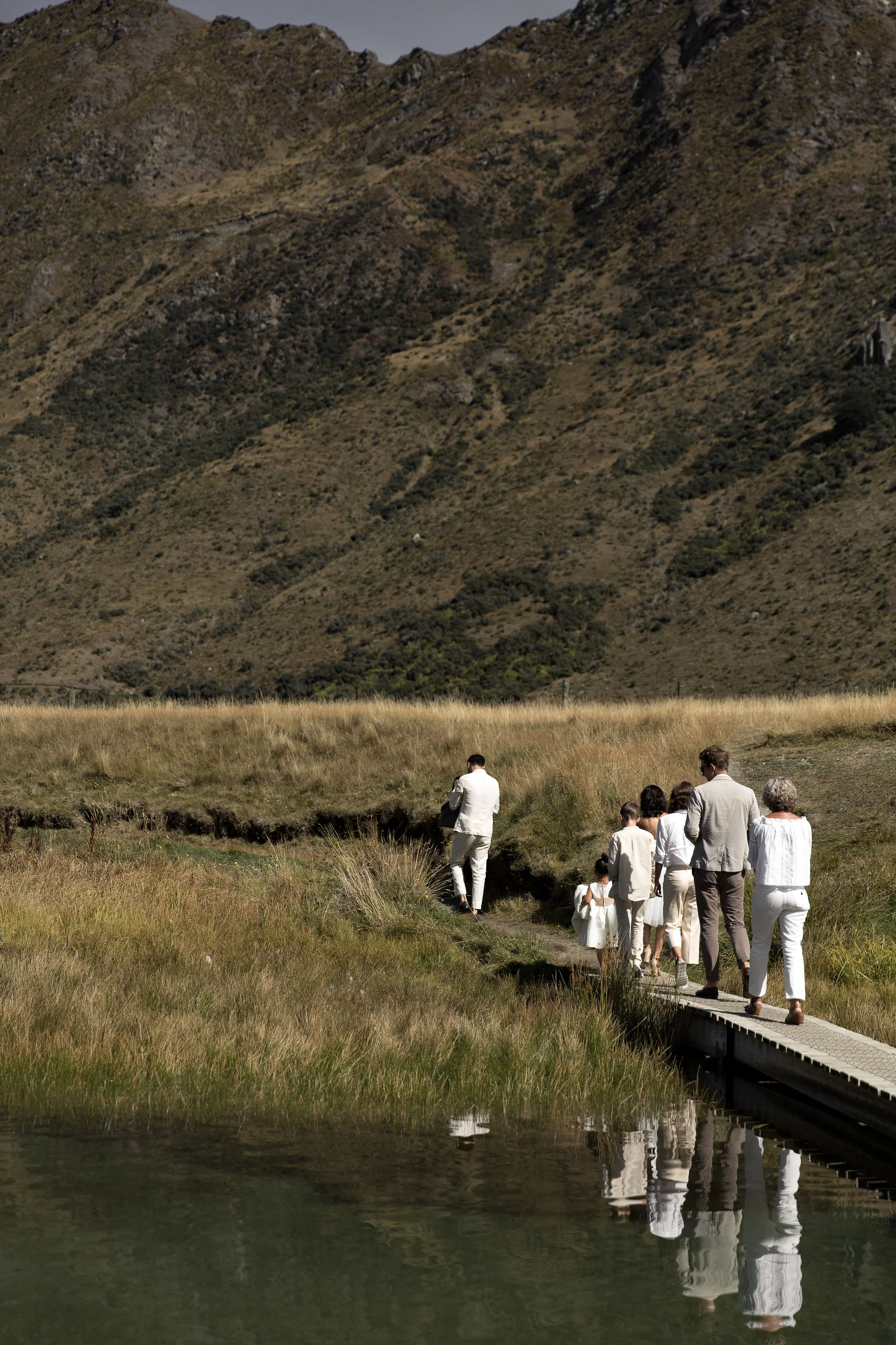 Group of people dressed in white walking on a narrow path across a grassy area with hills in the background, reflected in a body of water in the foreground.