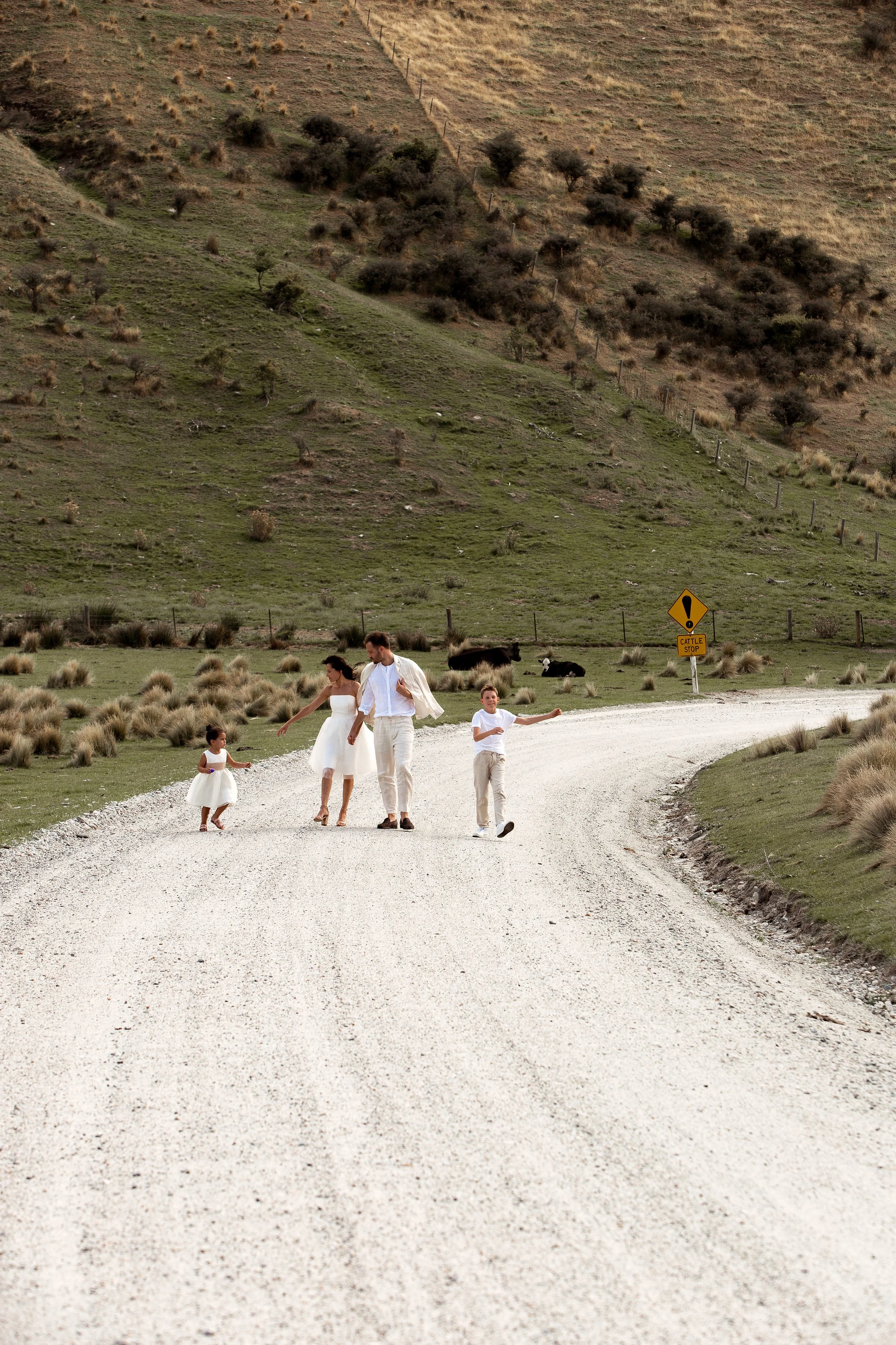 A family of four walking on a gravel road in a rural area with hills and cows in the background. The family is dressed in white and beige clothing.