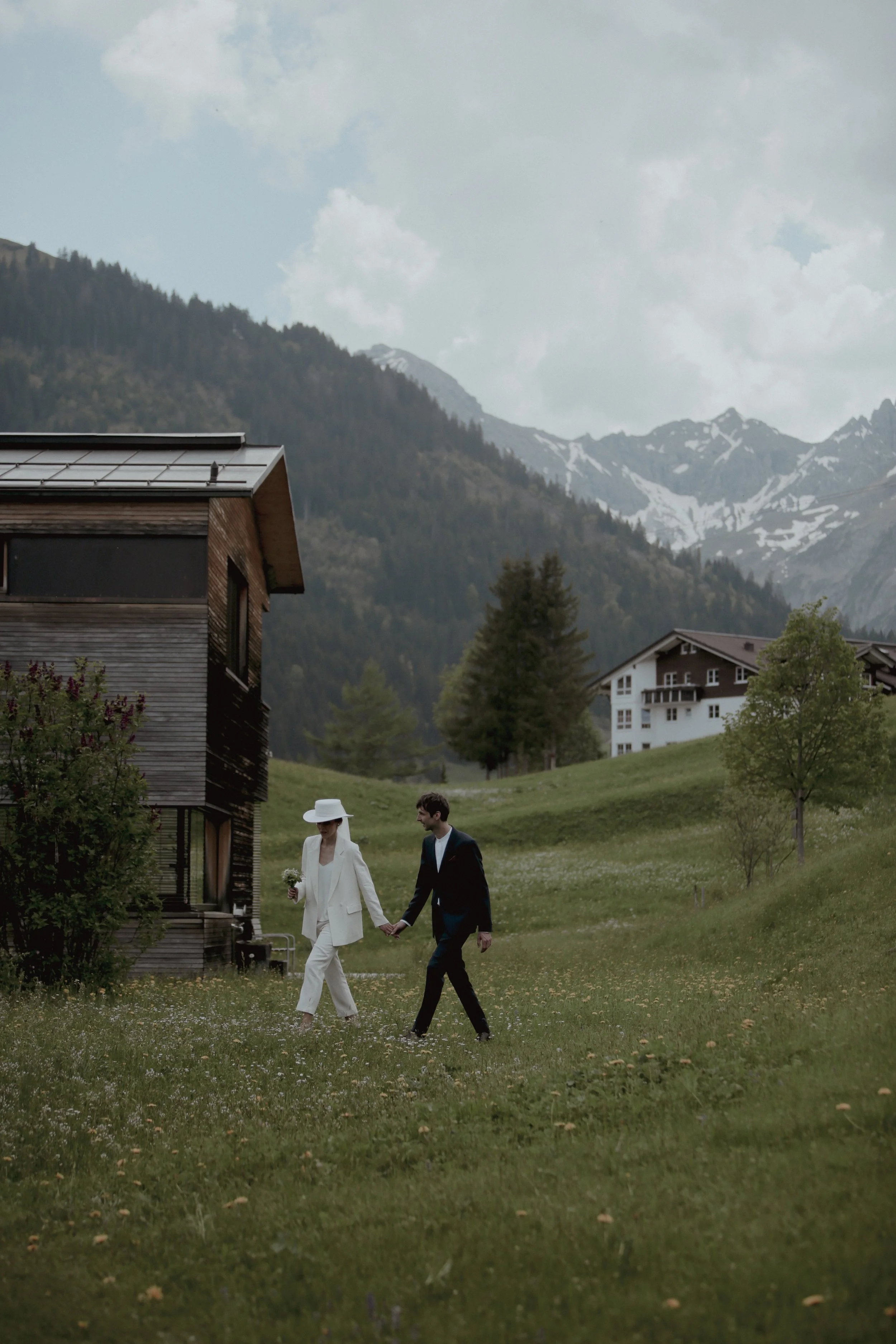 A couple dressed in formal wedding attire, walking hand in hand across a lush green field in a mountainous area with houses and snow-capped peaks in the background.