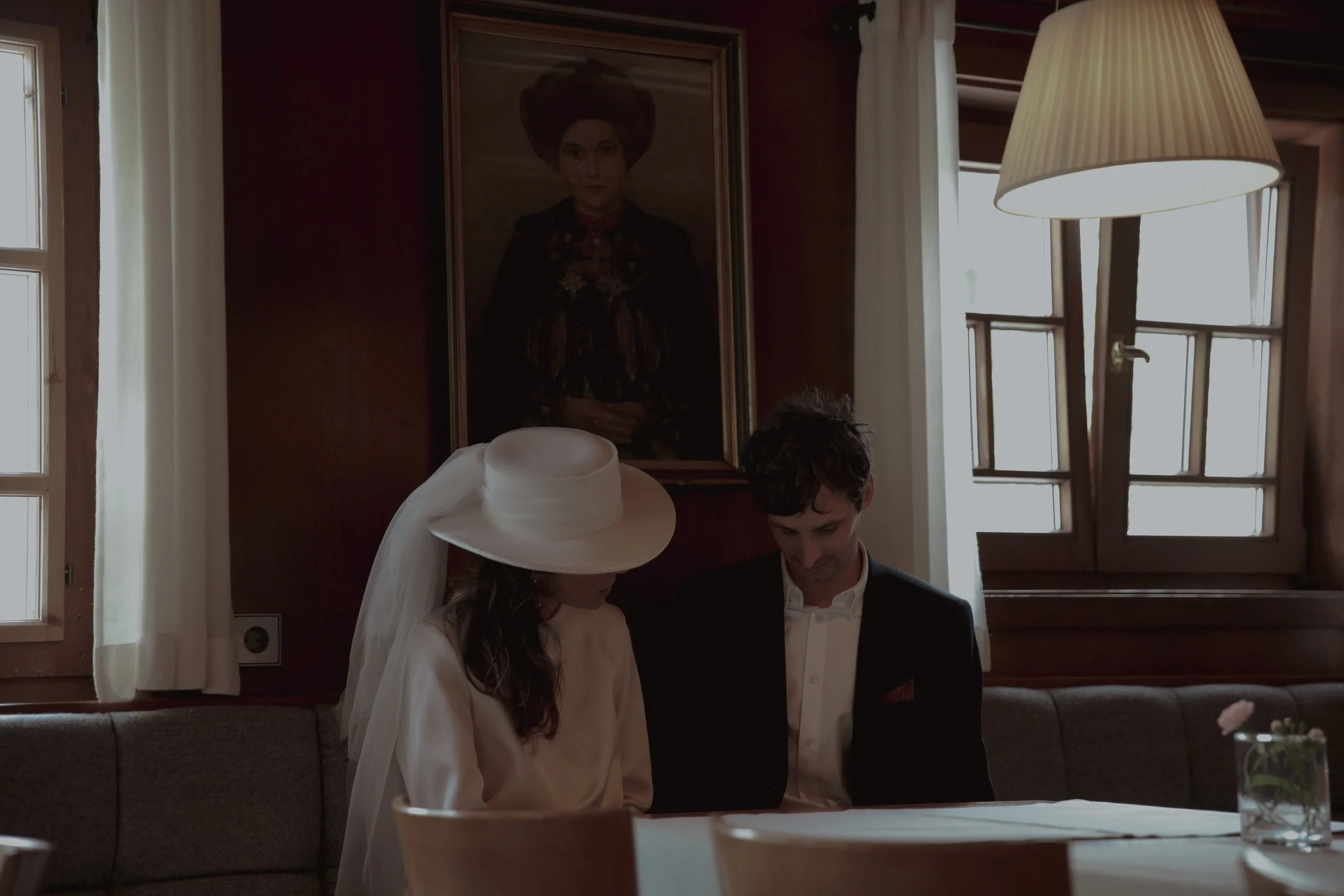 A bride in a white dress and large hat sits next to a groom in a dark suit, both looking down at something on the table. The setting is a cozy, wood-paneled room with a framed portrait of a woman hanging behind them, and a window with white curtains 