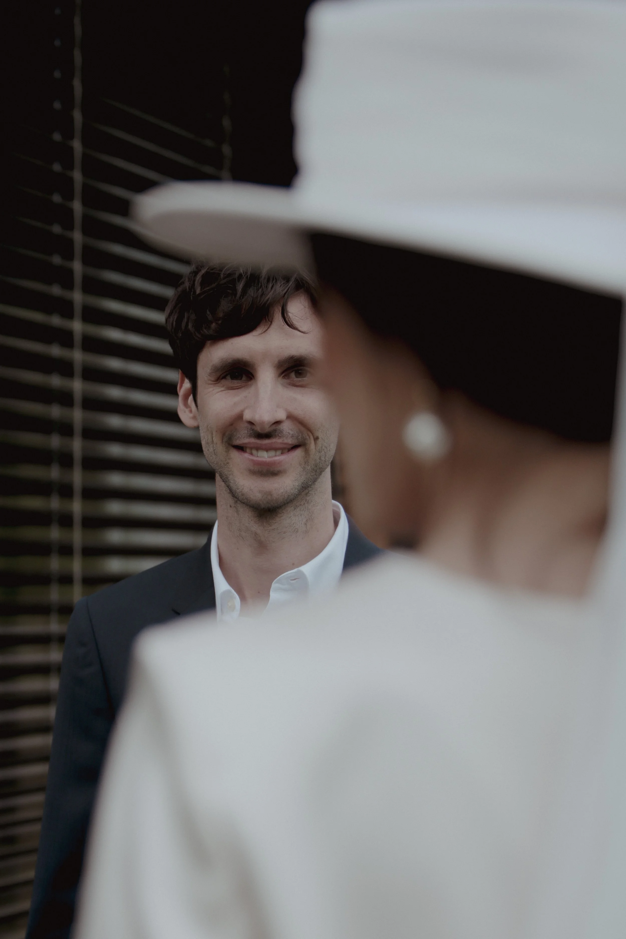 A man smiling, dressed in a dark suit and white shirt, standing outdoors in front of a slatted wall, engaged in conversation with a woman wearing a white hat and large pearl earrings, whose face is partially visible.