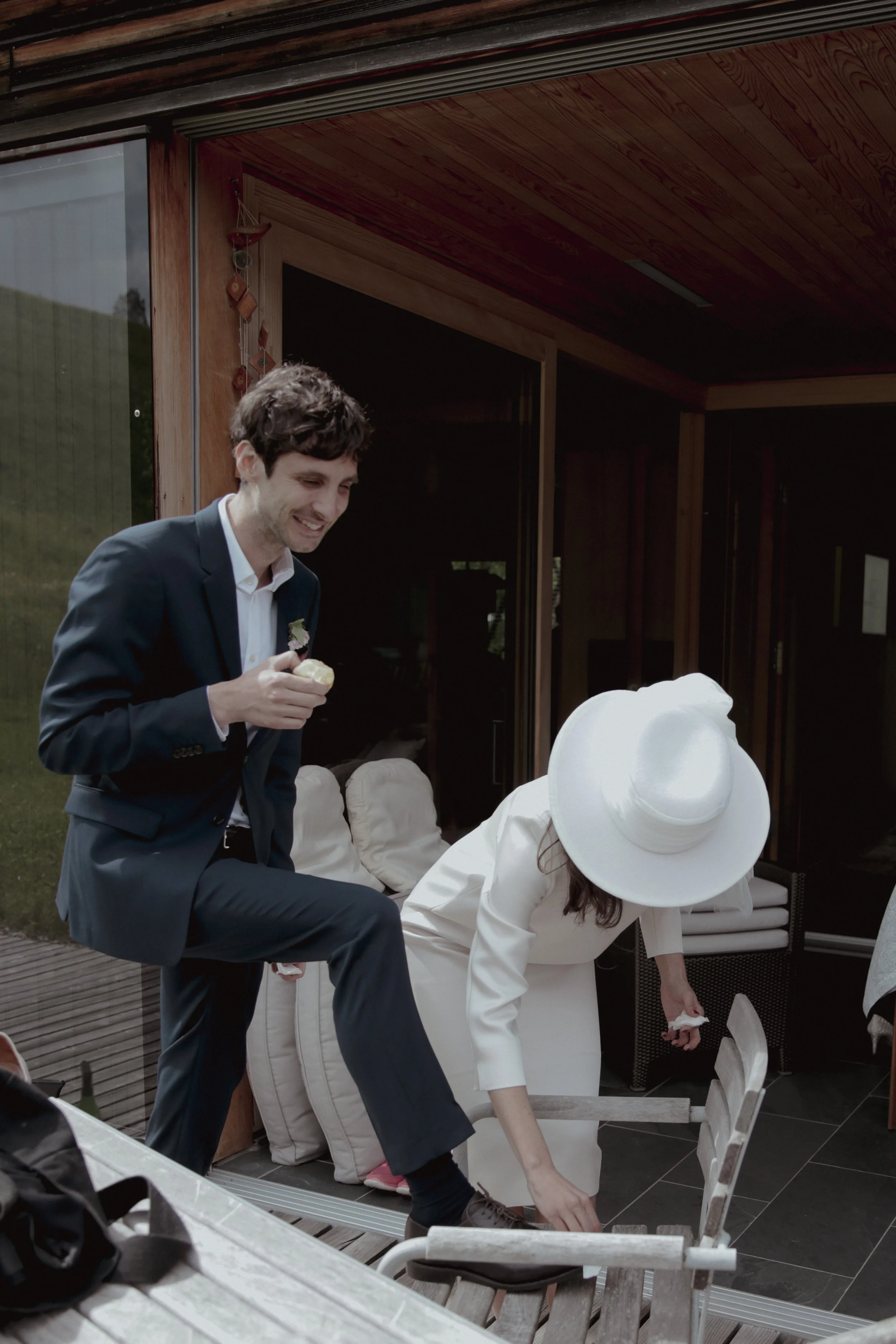 A man in a navy suit smiling and holding a cupcake, while a woman in white with a wide-brimmed hat bends down to adjust a chair on an outdoor patio.