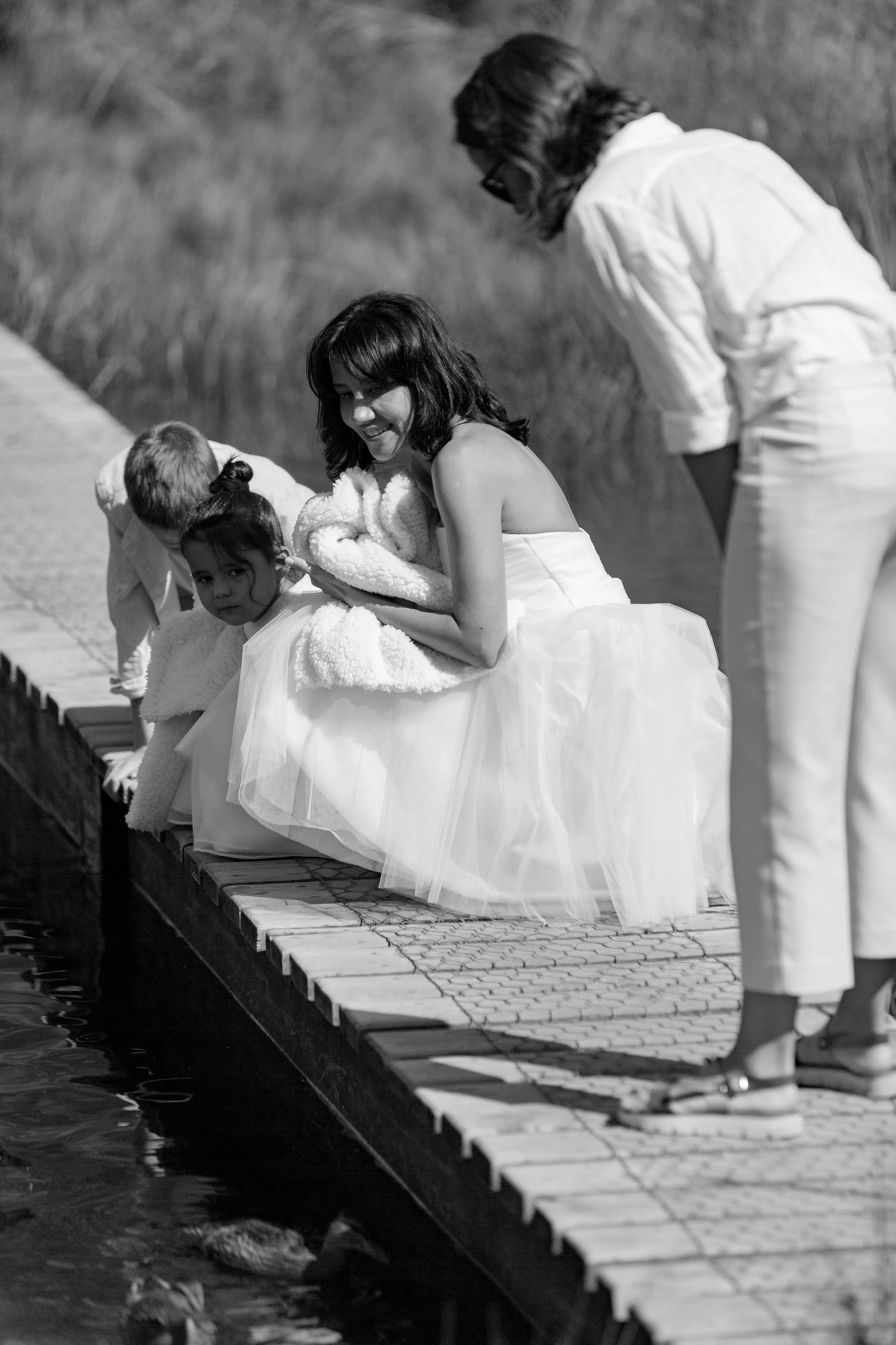 A woman and three children wearing white outfits sitting on a dock by the water, feeding ducks, in a black and white photograph.