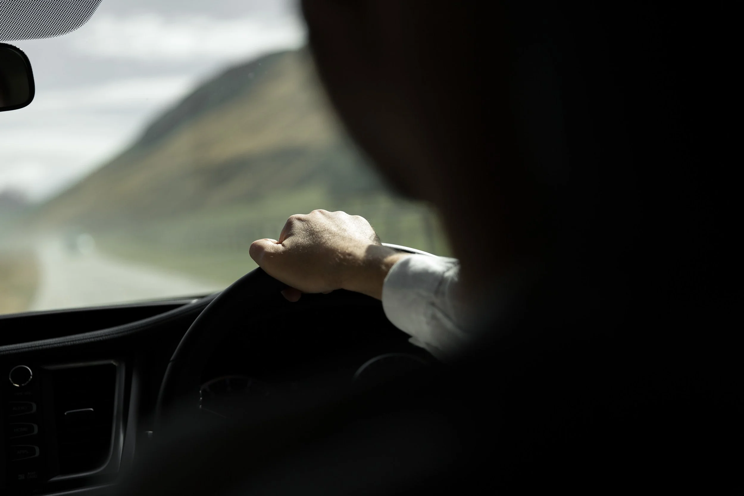 A person driving a vehicle with their left hand on the steering wheel, with a landscape of hills or mountains outside the windshield.